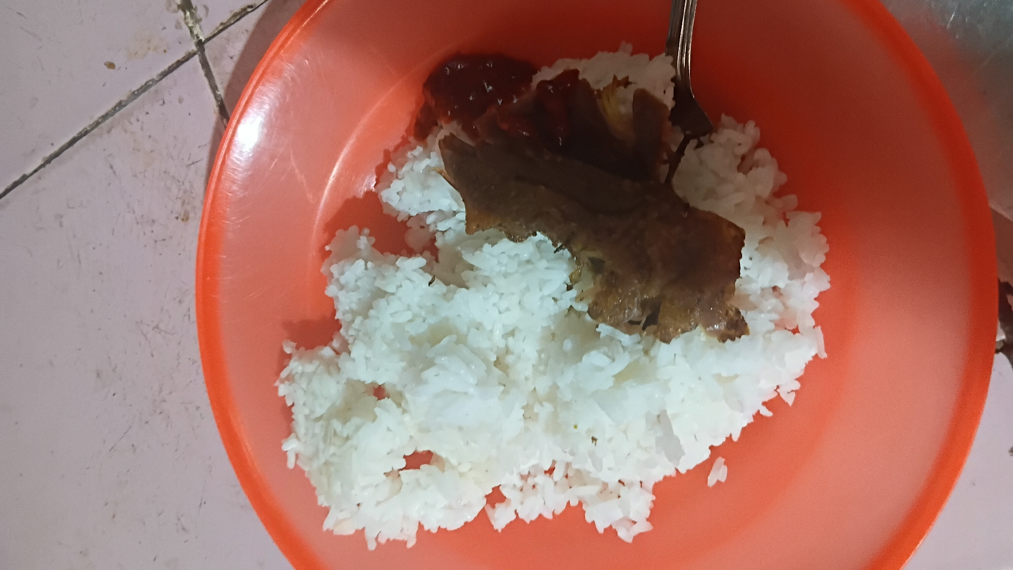 A top-down, close-up shot shows a portion of rice, meat, and sauce served on an orange plastic plate. The rice is white and fluffy, piled high on the left side of the plate. A piece of cooked meat, possibly chicken or beef, is placed on top of the rice, with some dark sauce underneath. To the right of the meat is a small portion of reddish-brown sauce, possibly sambal. A fork is visible on the right side of the plate, resting on the edge and partially touching the rice. The plate sits on a light pink, tiled surface with visible grout lines, suggesting it might be a table or countertop. The overall lighting is dim. The image location is not specified in the context provided.