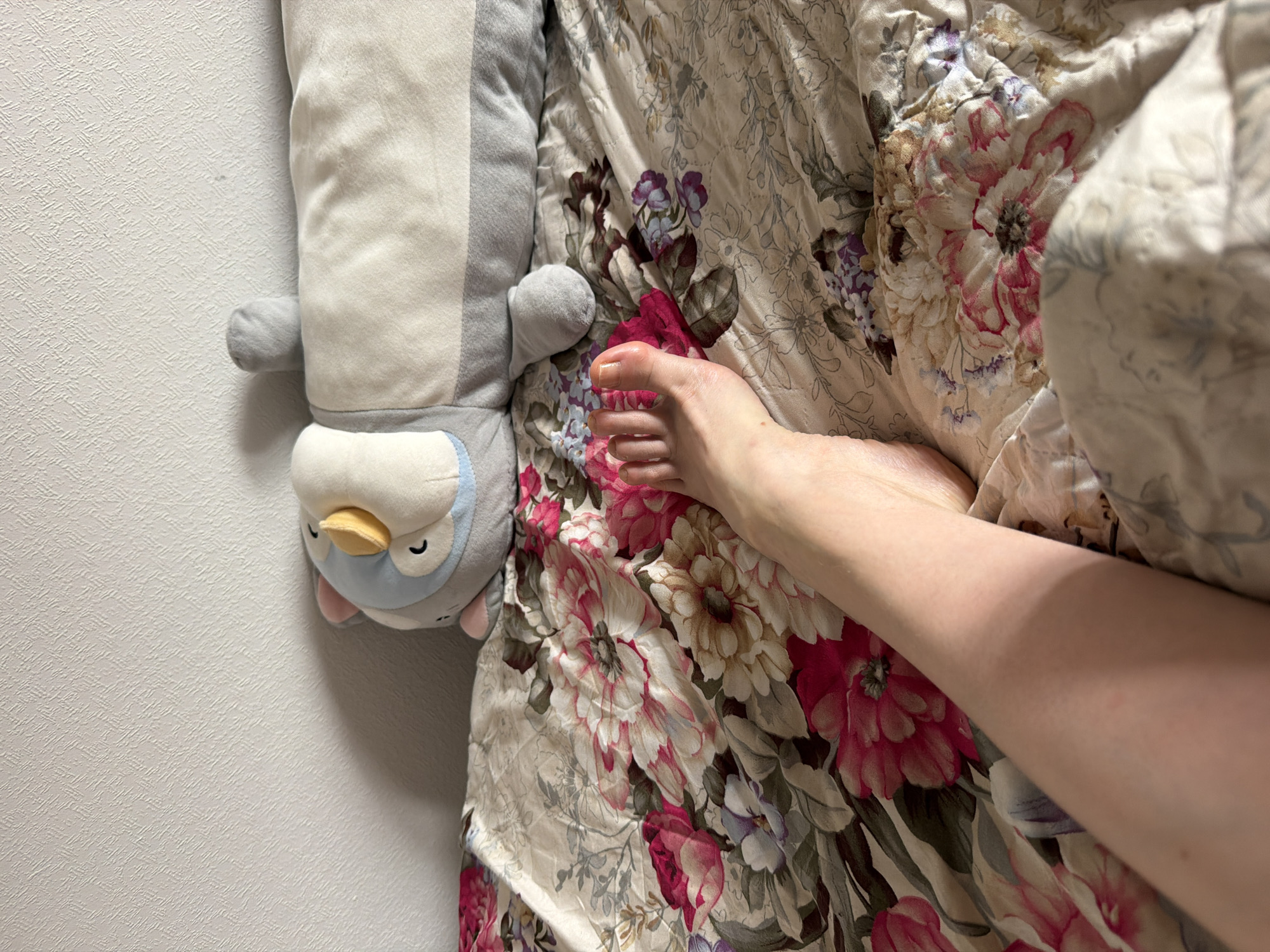 A plush penguin pillow lies on a floral patterned bedspread next to a pale skinned human leg and foot. The setting appears to be indoors, likely in a bedroom or living space. The background is a plain, light-colored wall. There is no indication of the location being Moscow, Russia.
