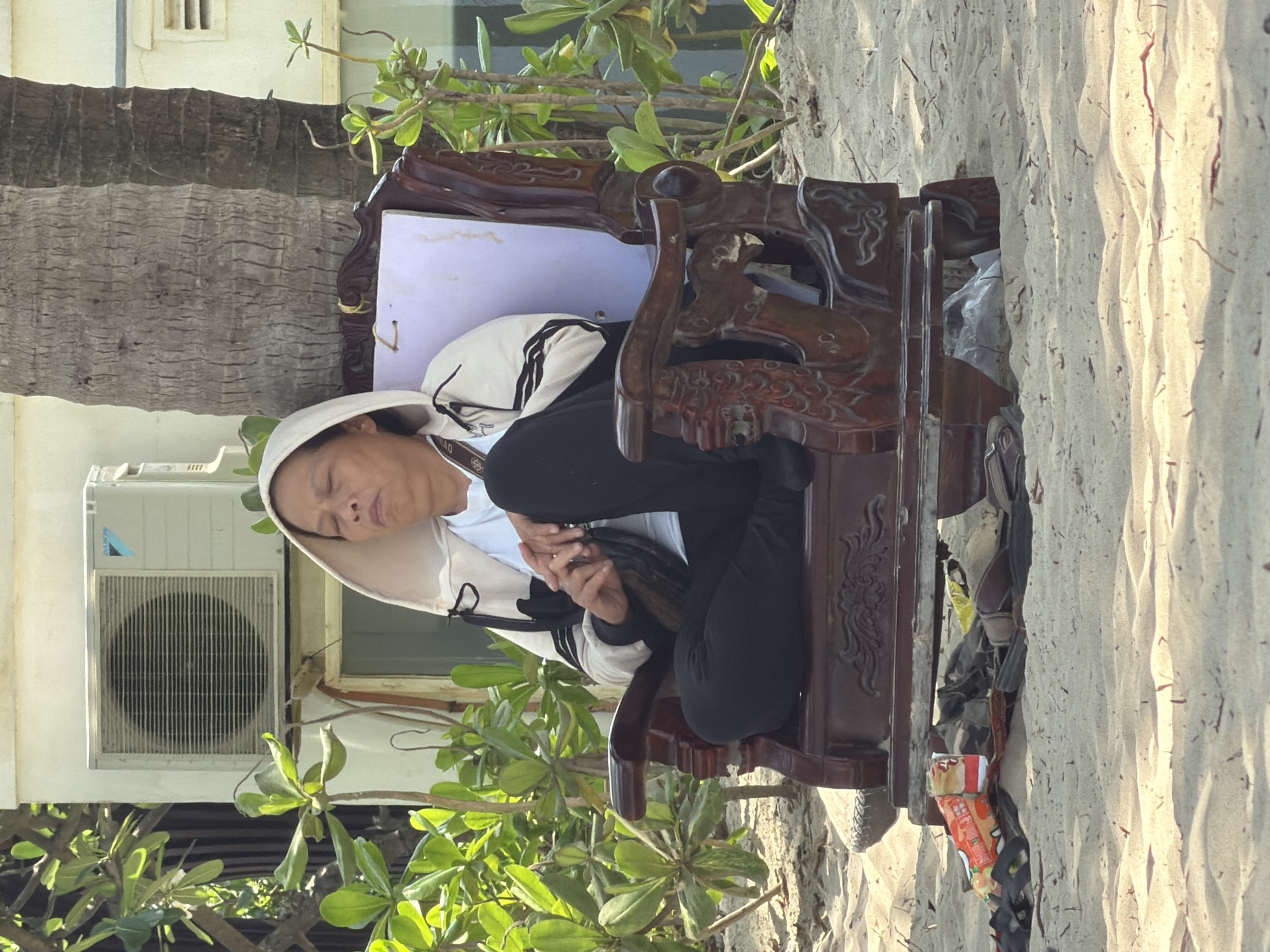 The image shows a person sleeping in a carved wooden chair on a sandy beach. The individual is wearing a white conical hat and a black jacket over a white shirt. The background includes tropical vegetation, a building with an air conditioning unit, and a palm tree. The sand appears to be on a beach in Đà Nẵng, Vietnam. Additional items are located under the chair and on the sand.