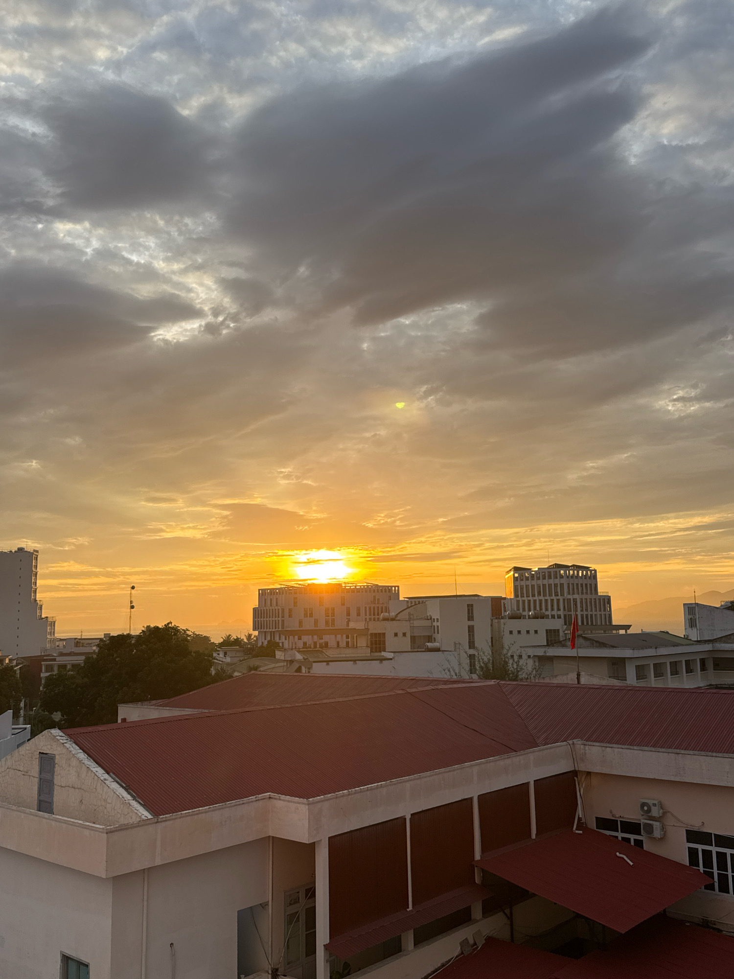 The image depicts a cityscape at sunset in Nha Trang, Vietnam. The sky is filled with dramatic, heavy clouds tinged with orange and yellow hues from the setting sun, which is visible as a bright orb just above the horizon, partially obscured by clouds. In the foreground, several buildings with distinctive red, sloped roofs are prominent. These buildings appear to be institutional or commercial, with some featuring white facades and modern architectural elements. Beyond the immediate foreground structures, a cluster of taller buildings, including a building with a unique, tiered facade and a flag atop it, rise into the sky. Trees are visible between the buildings, adding a touch of greenery to the urban landscape. In the distance, hazy mountains and a sliver of the sea are discernible. The overall scene captures a tranquil yet dynamic moment of a coastal city at dusk.
