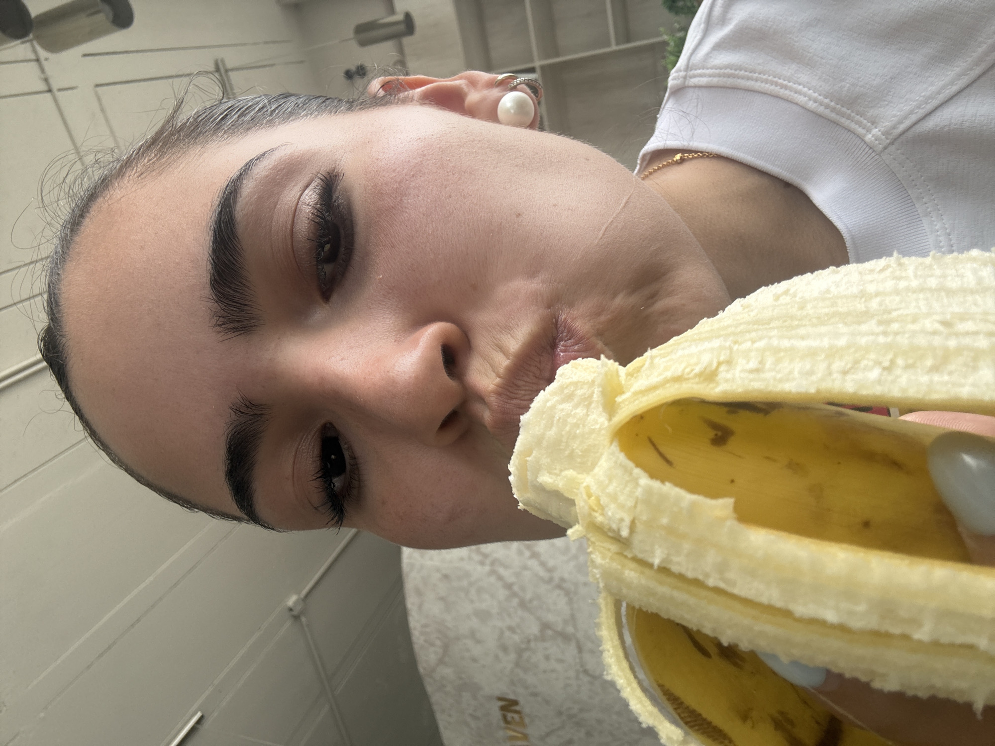 A close-up, angled shot depicts a woman holding a partially peeled banana close to her face, seemingly about to take a bite. The banana is yellow and has brown spots. Her skin is fair, and she has dark eyebrows and eyelashes. She is wearing a pearl earring and a gold chain necklace. The background appears to be an outdoor setting. The image is rotated counter-clockwise. In the lower left corner, a faint text "VEN" is visible. The location is unknown.
