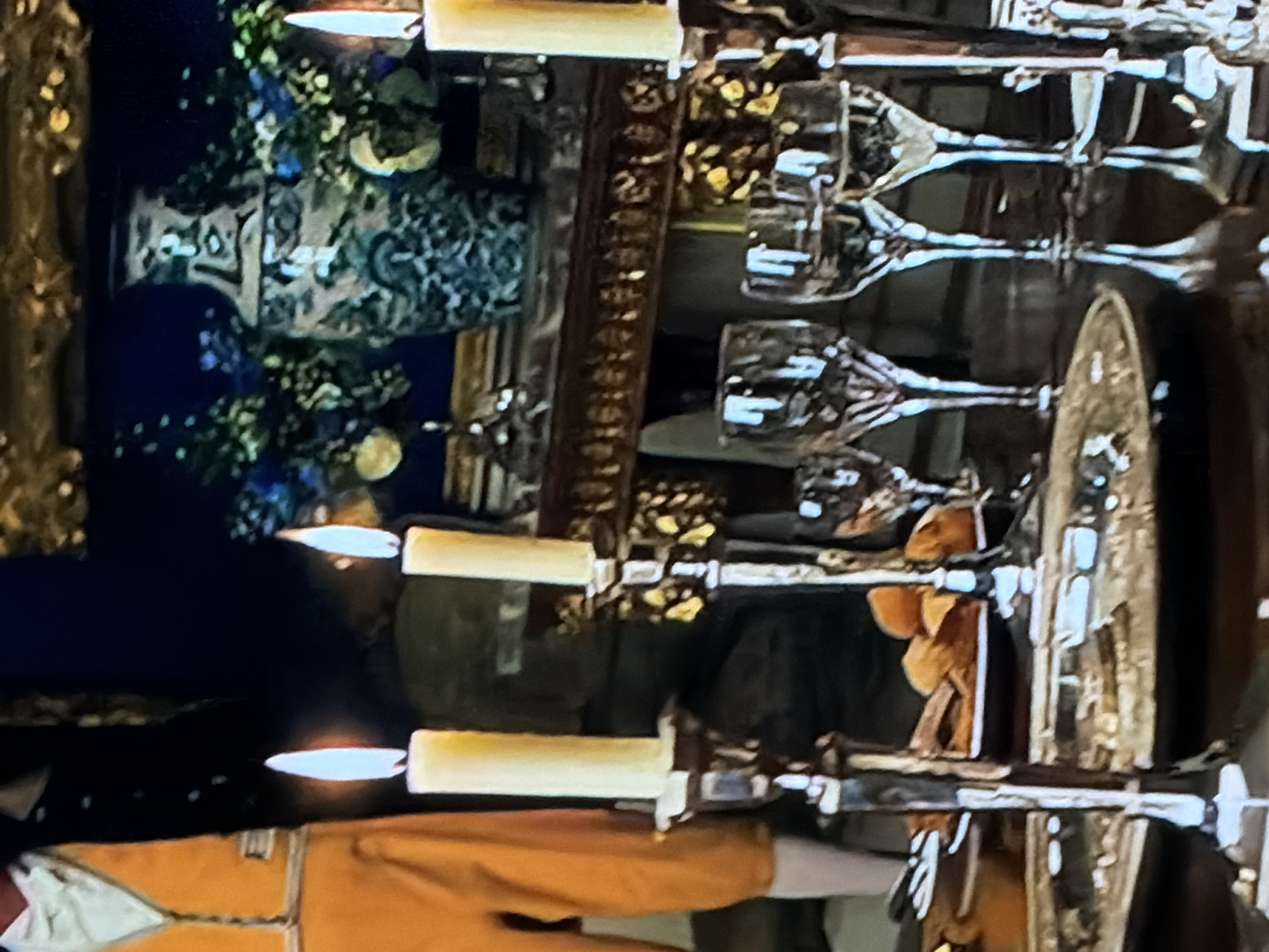 The image captures a lavish indoor scene, likely a banquet or formal gathering, set in what appears to be a historical or period-themed environment in Sofiivska Borshchahivka, Ukraine. The dominant feature is a long, ornate table set with numerous crystal glasses, gleaming candelabras holding lit candles, and gilded serving dishes. The candlelight creates a warm, opulent glow against a dim background, suggesting an evening event.
In the midground, a dark, intricately patterned vase or urn, adorned with green foliage and hints of blue flowers, serves as a centerpiece. A highly decorative, dark brown element, possibly a table runner or part of the table itself, runs along the center, showcasing repeating, complex designs. The reflective surface of the table clearly shows inverted reflections of the glassware and candles.
Towards the foreground and lower left of the frame, a person is visible, dressed in what appears to be period costume. They wear an orange or golden-hued jacket with white trim on the sleeves and collar, possibly over a white undershirt or cravat, suggesting attire from an earlier century. Their stance indicates they are standing beside the table.
The scene conveys a sense of grandeur and formality, characteristic of a significant celebration or a historical reenactment. No specific activity beyond the setting of the table is captured, but the prepared state implies an event is underway or about to begin. There is no visible text in the image.