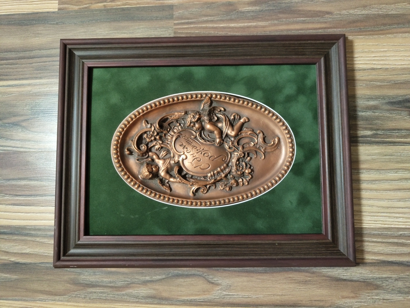 A framed oval plaque with a copper-colored relief of two cherubs and floral elements is displayed on a green velvet background. The plaque has the inscription "С Днем рождения" (Happy Birthday) in Cyrillic script. The frame is dark brown wood and is set against a wooden plank background. The image is taken from a slightly elevated angle, showing the top and left sides of the frame. No specific location in Chișinău, Moldova can be determined from the image.