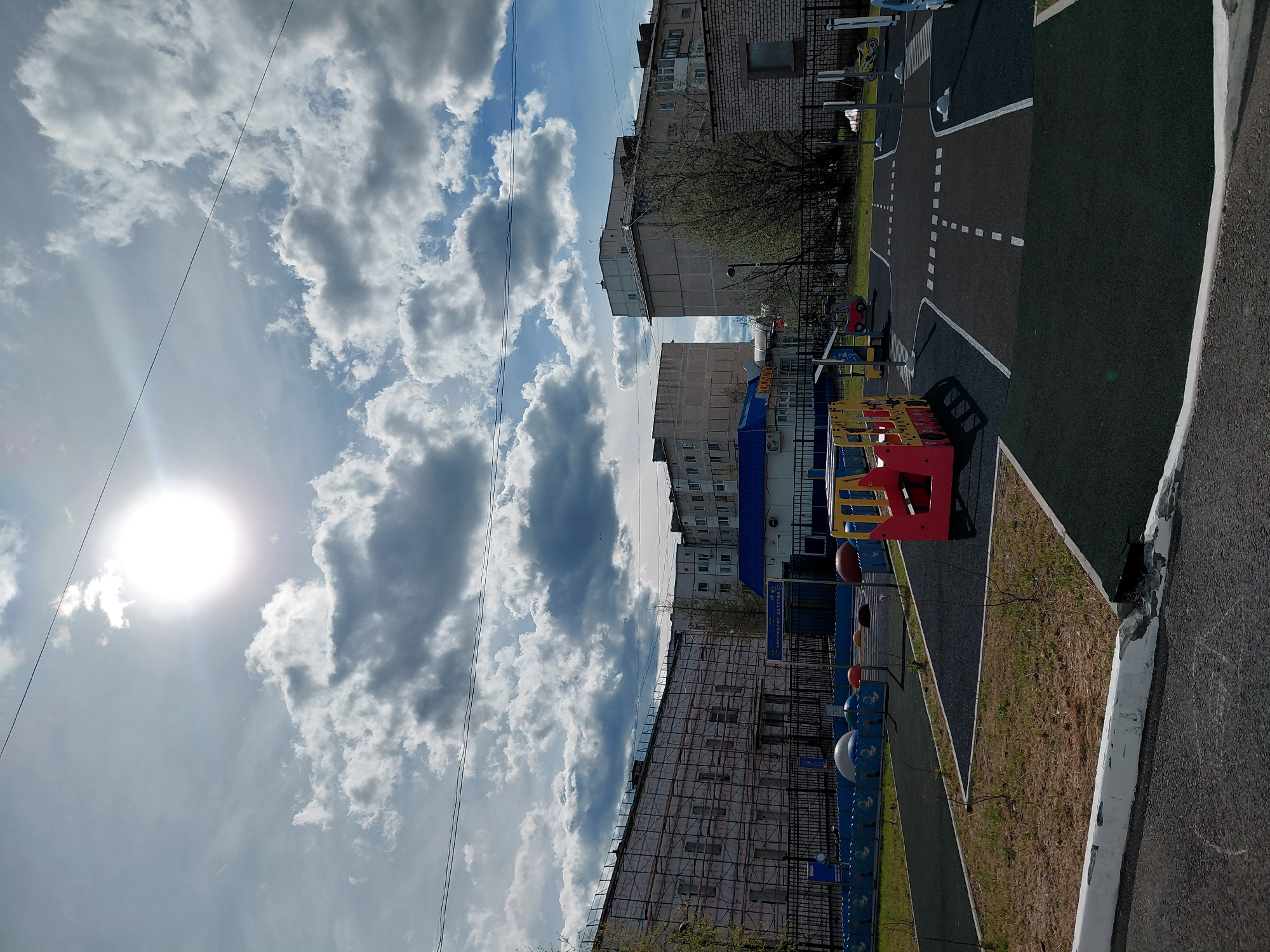 The image captures an outdoor children's playground or traffic education area in Nadym, Russia, during a bright, partially cloudy day. The sun, visible in the upper left, casts a strong glare. The foreground features a dark, rubberized surface designed to resemble a road, complete with white dashed and solid lines. A red and yellow play structure, possibly a small bus or car, and miniature traffic lights are placed on the left side of this simulated road. A white, somewhat cracked curb runs along the right edge of the road.
In the background, several multi-story residential buildings are visible under the sky. Their facades vary from brick to grey concrete, and one building appears to have scaffolding or a metal structure on its side. Leafless tree branches are scattered between the buildings and the play area, suggesting early spring or late autumn. A blue sign prominently displays the Cyrillic text "КРОССИНГ ДЕТСТВА," which translates to "CHILDHOOD CROSSING," indicating the purpose of the facility. The sky is bright blue with numerous fluffy white and grey clouds, and a utility wire crosses the upper portion of the scene. No people are present, and no specific activity is currently taking place.
