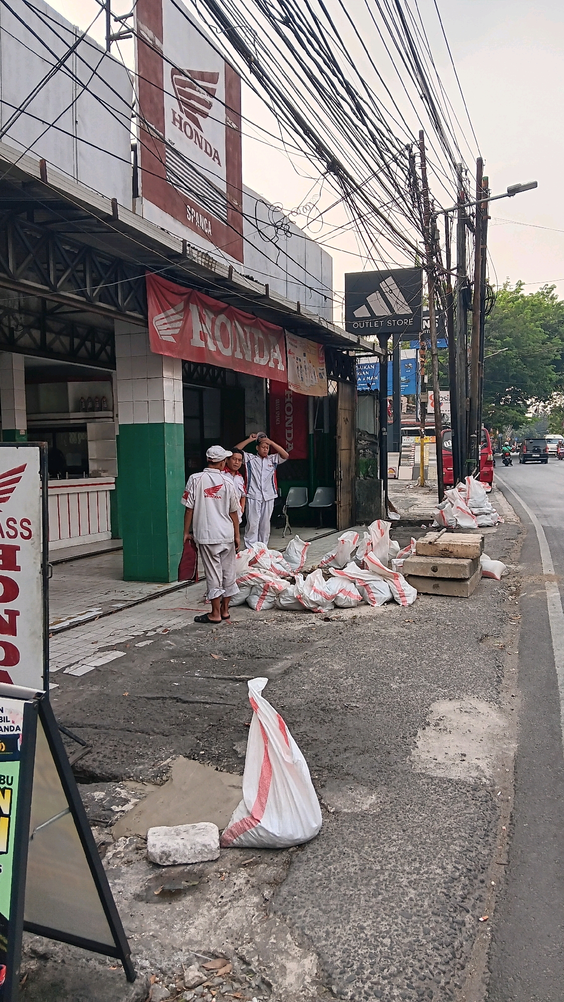 The image depicts a street scene in South Jakarta, Indonesia, in front of a Honda outlet. A large red banner with the Honda logo and "HONDA" text hangs prominently above the entrance. To the right of the entrance, an Adidas outlet store sign is visible. In front of the Honda outlet, three individuals, two appearing to be male and one female, are standing on the sidewalk. All three are wearing white uniforms with "HONDA" logos. One individual is adjusting their headwear, while another appears to be talking. A pile of white sacks with red stripes are stacked on the sidewalk next to the individuals. The sidewalk is uneven and cracked, with some debris and a single white brick present. A street runs alongside the sidewalk, with a few vehicles, including motorcycles and cars, visible in the distance. The sky is overcast, suggesting it might be late afternoon or early morning. The overall scene conveys a sense of everyday commerce and street activity in an urban environment.