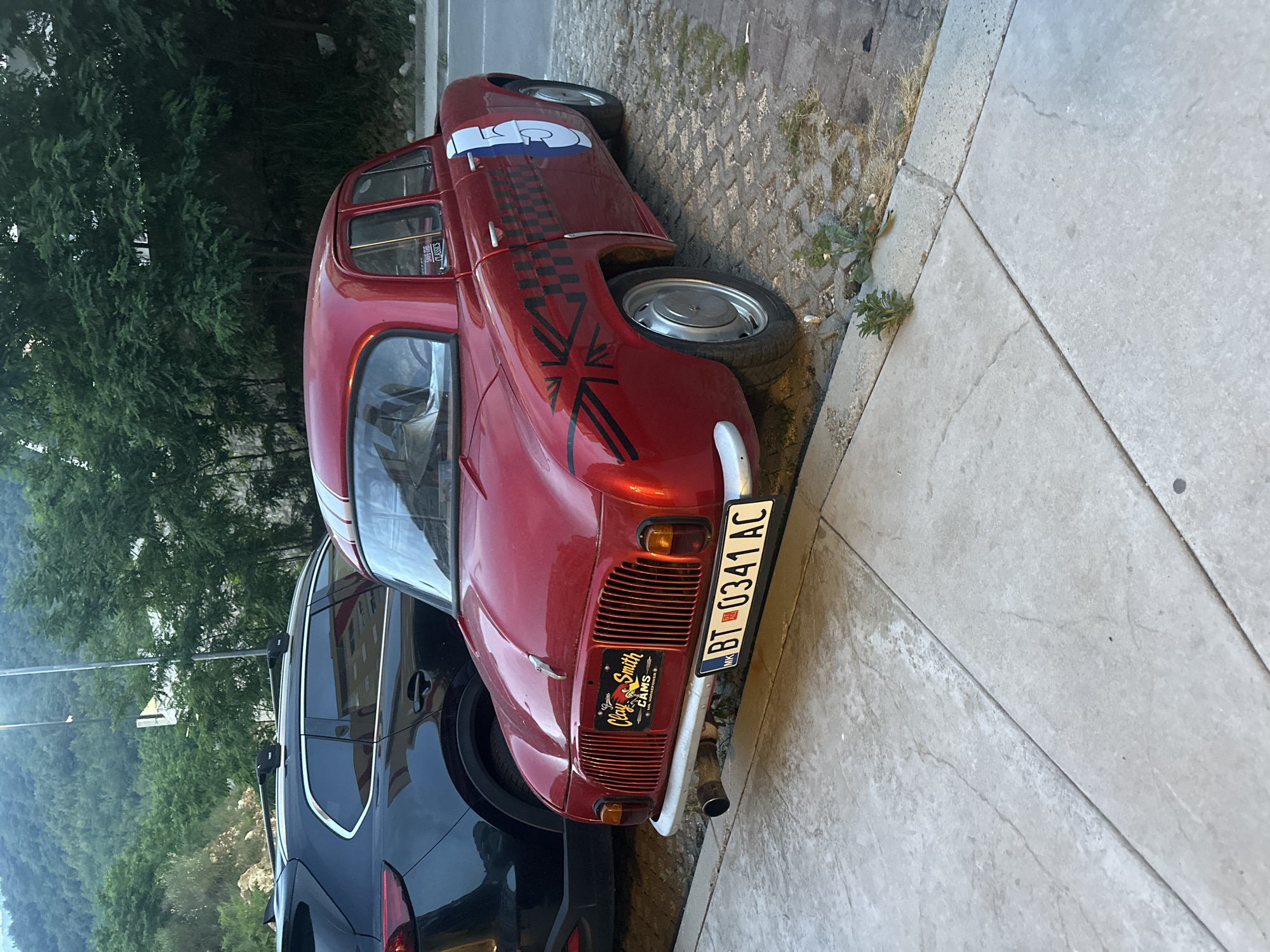 A vibrant red, customized vintage car, resembling an older European model such as a Renault 4, is the central subject of this image. The car is parked on a cobblestone-like surface, next to a modern, dark-colored SUV partially visible on its left. On its right, there is a lighter concrete sidewalk with some weeds growing in the cracks. The car features several unique decals: a prominent white racing number "5" on its side, a black and white checkered pattern stretching from the front fender to the door, and a stylized black Union Jack flag graphic on the lower front fender. A white racing stripe runs along the roof.
The car's rear shows a chrome bumper and a distinctive grille design. Its license plate, "BT 0341 AC," is a Macedonian plate with the country code "MK." Below the license plate, a smaller plaque or sticker reads "Cloy & Smith CAMS" with the word "Classic" beneath it. Another sticker, visible on the rear side window, says "SUPER RIDE CLASSICS." The exhaust pipe is visible protruding from the rear.
In the background, lush green trees and what appears to be a mountainous or hilly landscape dominate, suggesting an outdoor setting in a natural area, consistent with Boreti, Montenegro. The lighting indicates it might be late afternoon or early evening. No people are visible, and the cars are stationary.
