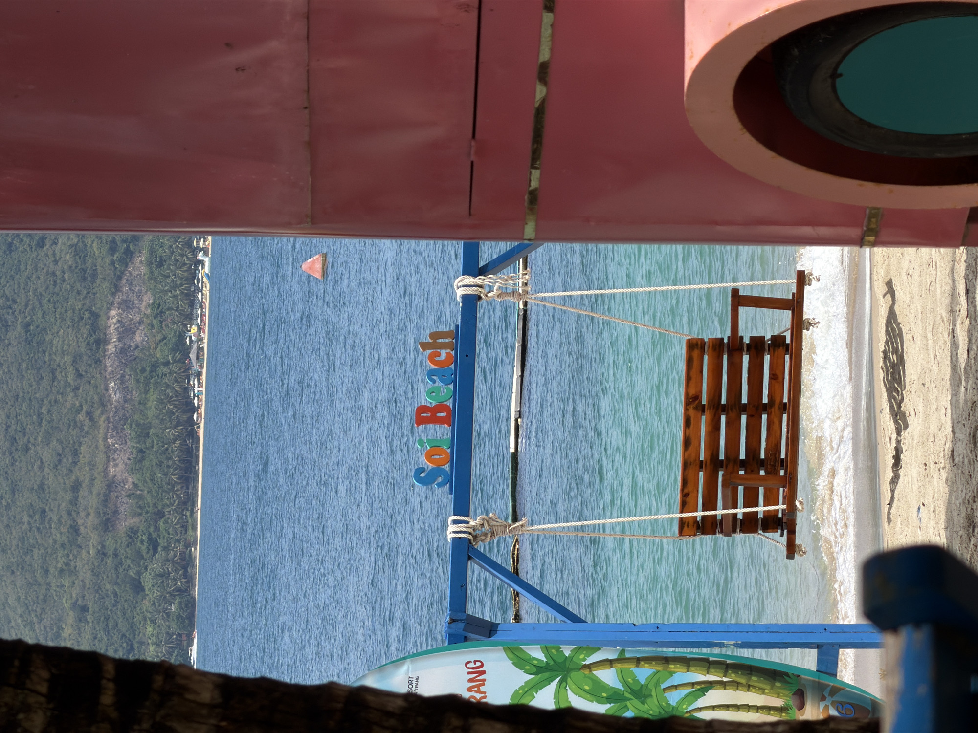 A topsy-turvy outdoor scene shows a swing hanging over a beach, likely in Nam Nha Trang Ward, Vietnam, based on the "Soi Beach Trang" sign. The swing, made of wood and suspended by ropes, appears over the sandy shore where waves meet the land. Above the swing and beach, the image is framed by architectural elements: a pink, possibly metal structure with a circular opening containing a teal-colored insert. Below the swing, a blue frame supports a surfboard with a palm tree design. In the distance, a densely forested area stretches along the coast, next to buildings along the shoreline. A small orange buoy floats in the water.