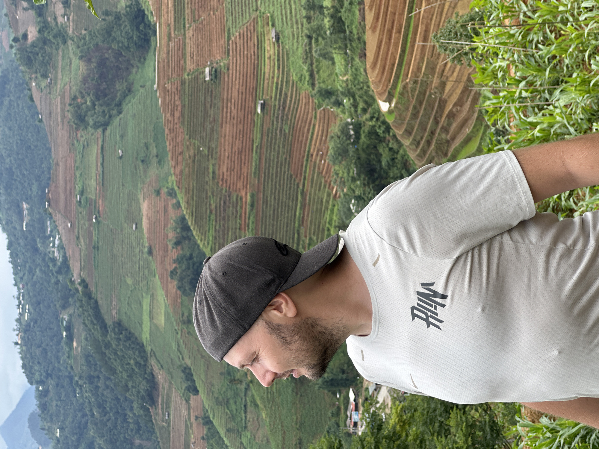 A male individual is depicted in a mid-shot, positioned in the right foreground and facing left in profile. He has a beard and is wearing a dark grey baseball cap backward, and a light grey short-sleeved athletic shirt with a stylized black "RUN" logo on the left chest. He is looking towards the left with a slight smile. The background reveals a panoramic view of layered rice terraces covering undulating mountainous terrain. The terraces exhibit varying hues of green, indicative of cultivation, and reddish-brown, suggesting fallow or tilled land. Small, scattered structures are visible across the hillsides. Lush green vegetation, including what appears to be corn stalks, occupies the immediate foreground on the right. The scene is set in the mountainous region of Dế Xu Phình, Vietnam, under an overcast sky.