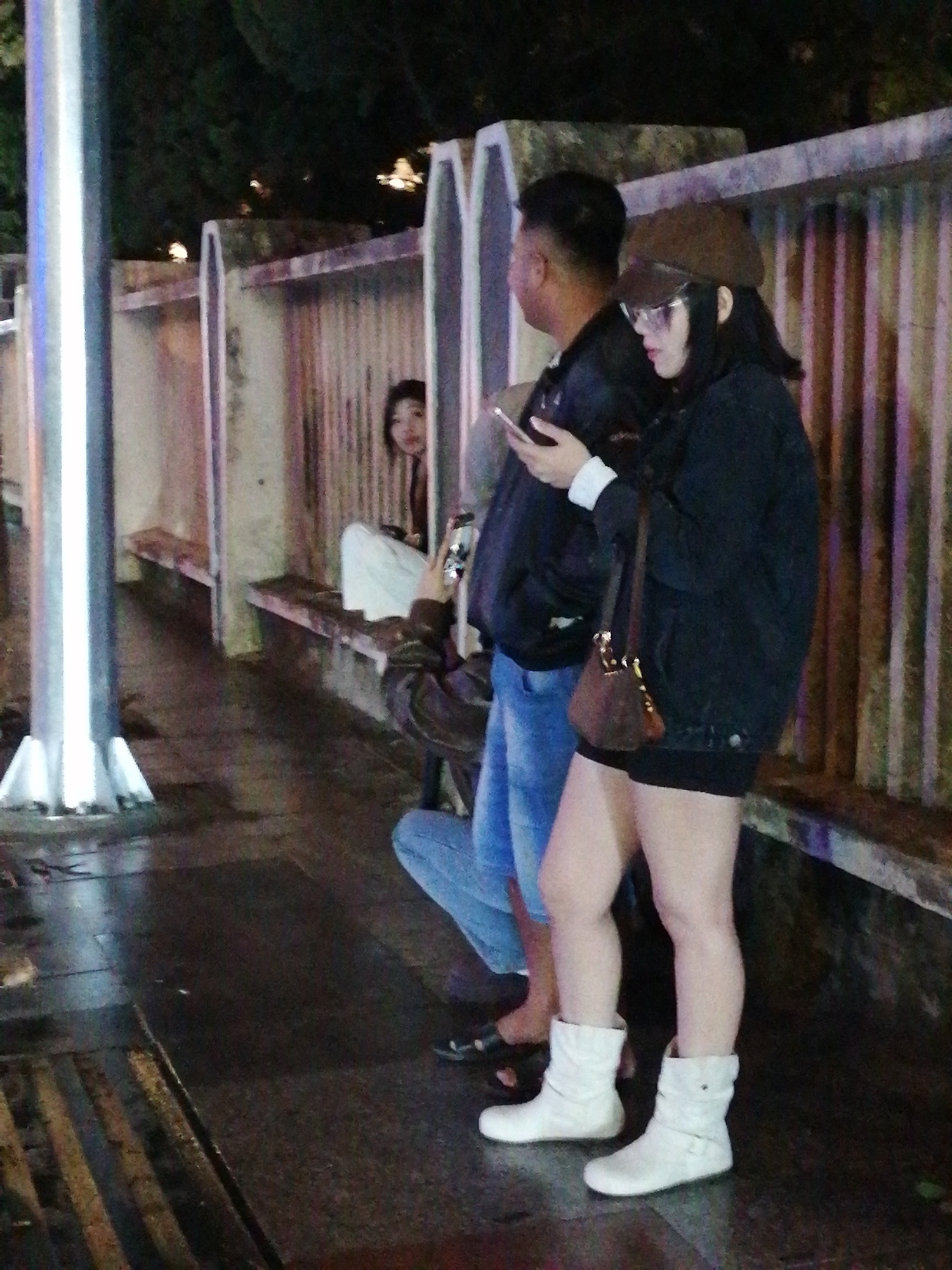 The image depicts a nighttime scene in Da Lat, Vietnam, specifically near a covered seating area with benches. A young woman, wearing a brown cap, sunglasses, and a black jacket over a white shirt, stands to the right, holding a phone. She has a small brown purse slung across her shoulder. To her left, a man in a black jacket and blue jeans is also visible. Further back, another person is seated, partially obscured. The ground appears wet, suggesting it might have rained recently. In the background, a fence-like structure runs along the seating area, and trees are dimly visible beyond it. A tall, cylindrical pole with a reflective surface stands on the left side of the frame.