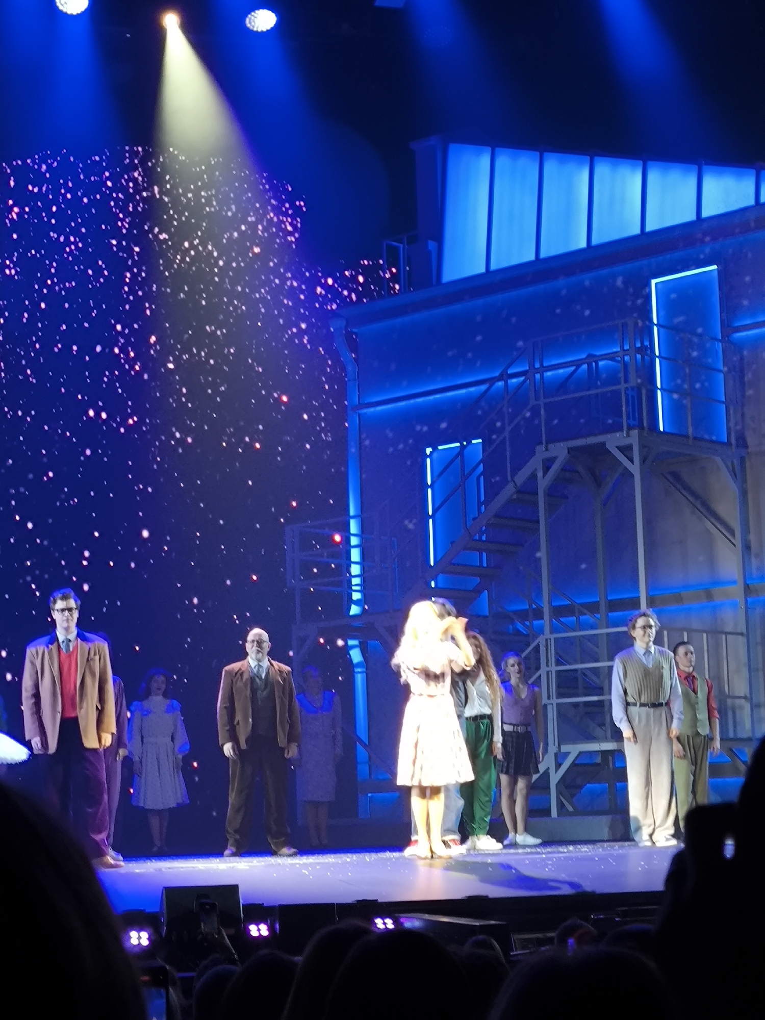 A stage production is underway, set against a dark blue backdrop illuminated by a spotlight and a scattering of bright white and pink lights resembling confetti or stars. A blue, multi-level structure with stairs and railings dominates the right side of the stage. A group of people, likely actors, are positioned on the stage in a line.
From left to right:
A man in a brown jacket, red sweater, and dark pants stands with glasses.
A woman in a white dress is next to him.
A man in a brown suit and waistcoat stands with a bald head and glasses.
A woman in a light-colored dress is partially visible.
A woman in a purple top and black skirt stands next to her.
A man in a light brown vest over a blue shirt and tan pants stands.
A man in a red vest over a white shirt and tan pants stands.
The lighting is theatrical, with bright blue washes on the set and a focused white spotlight. The audience is visible in the foreground, with some holding up phones to record. The location is an unknown theatrical venue.