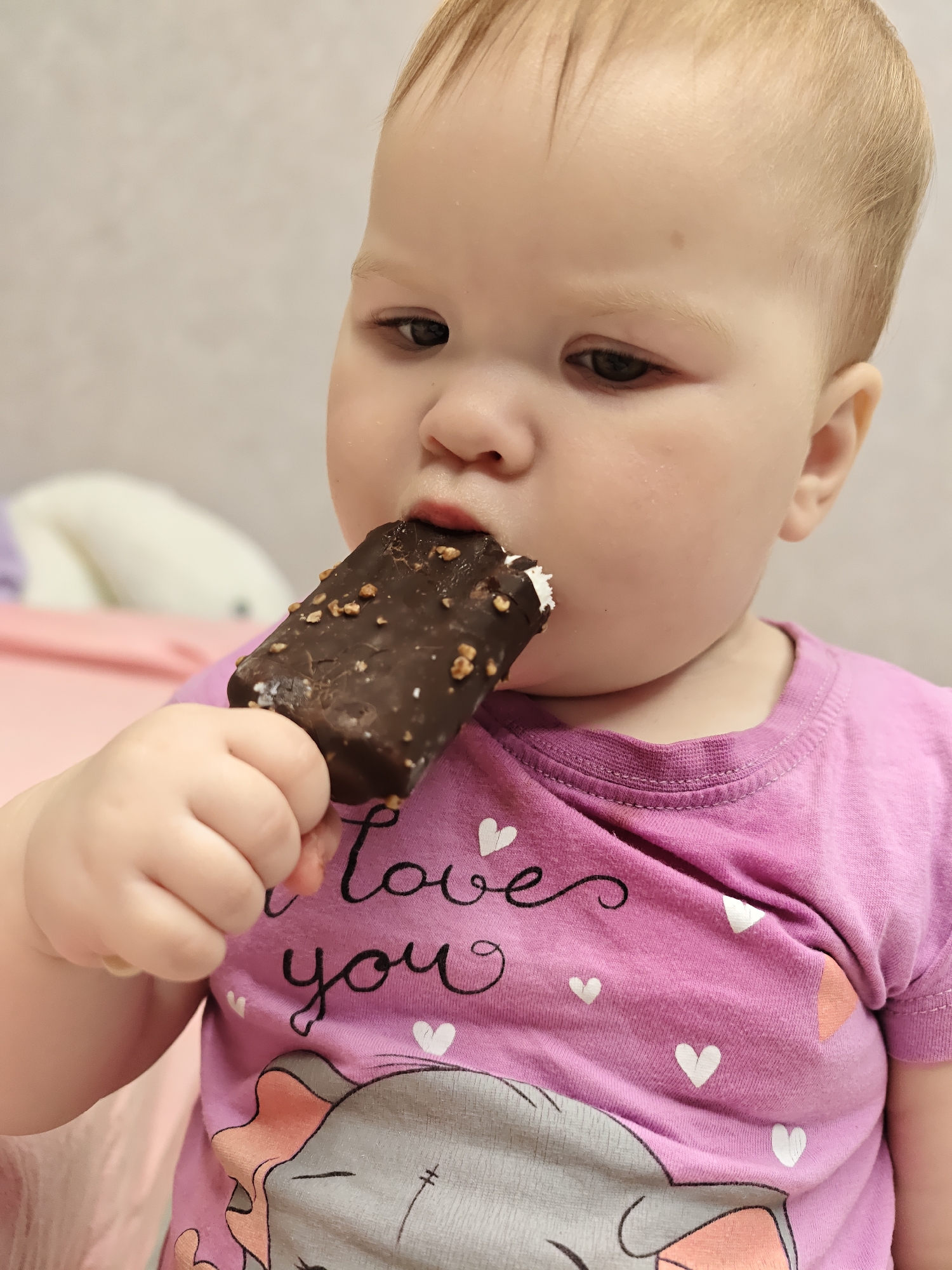 An indoor, close-up image depicts a fair-skinned baby or young toddler with light-colored hair, holding and biting into a chocolate-covered ice cream bar with visible nut pieces. The toddler's left hand grasps the ice cream, exposing a light-colored interior where a bite has been taken. The individual wears a fuchsia-colored, short-sleeved t-shirt featuring a design of white hearts, the phrase "Love you" in black cursive script, and a partial graphic of a gray and peach elephant head. The background is softly blurred, showing light-colored soft furnishings and a pink surface, consistent with a domestic environment. This scene is located in the city of Chaikovsky, Russia.
