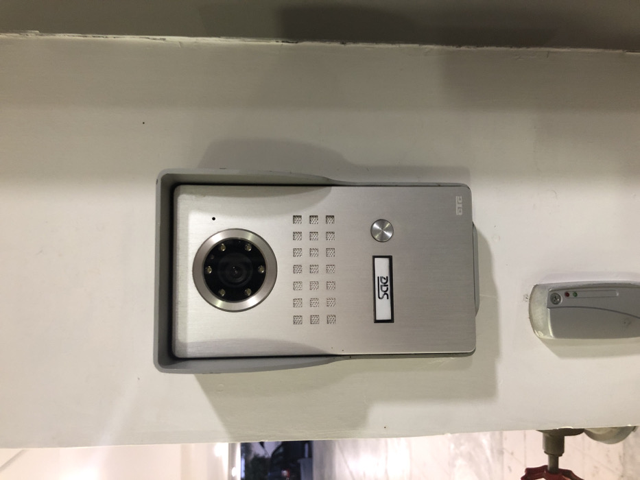 The image displays an outdoor intercom system and an access control unit mounted on a white wall or pillar. The primary subject is a rectangular, silver-colored intercom unit crafted from brushed metal. On its left side, a prominent circular camera lens is visible, surrounded by several small, dark circles that appear to be infrared (IR) LEDs. To the right of the camera, two grilles with square perforations are present, likely serving as a speaker and microphone. Further to the right, a circular silver button, presumably a call button, is positioned. Below this button, a rectangular slot contains a vertically oriented nameplate or display reading "DDS". In the top right corner of the intercom unit, a small logo or text resembling "DTG" or "DIG" can be discerned.
To the immediate right of the intercom unit, a smaller, oval-shaped greyish-silver electronic device is also mounted on the wall. This unit, possibly a card reader or access sensor, features two small indicator lights (one red, one green) on its left side. The background is a plain white wall or building exterior, with subtle shadows cast by the devices. The bottom edge of the original image shows a blurred background, possibly revealing reflections and part of a red pipe or valve. No people are visible, and no specific activity is occurring. The scene suggests an entrance area of a building in Moschato, Greece, under ambient or daytime lighting conditions.