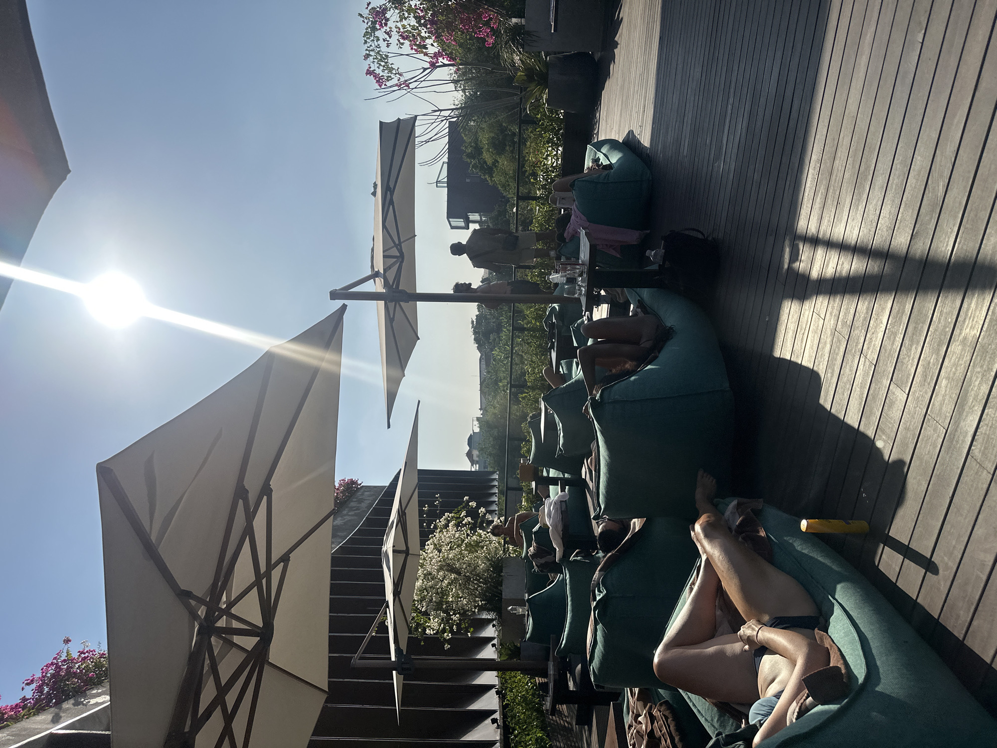 The image shows an outdoor rooftop seating area with several people relaxing on large, teal-colored beanbag chairs. The area is shaded by large white umbrellas, and the sun is shining brightly, casting long shadows on the wooden deck. In the background, there are buildings and lush greenery, suggesting an urban or resort setting. Some individuals are wearing swimwear, and there are tables with drinks and personal items. Two men are standing and conversing near the railing. A bottle of sunscreen is visible on the deck in the foreground. The overall atmosphere appears to be one of leisure and relaxation.