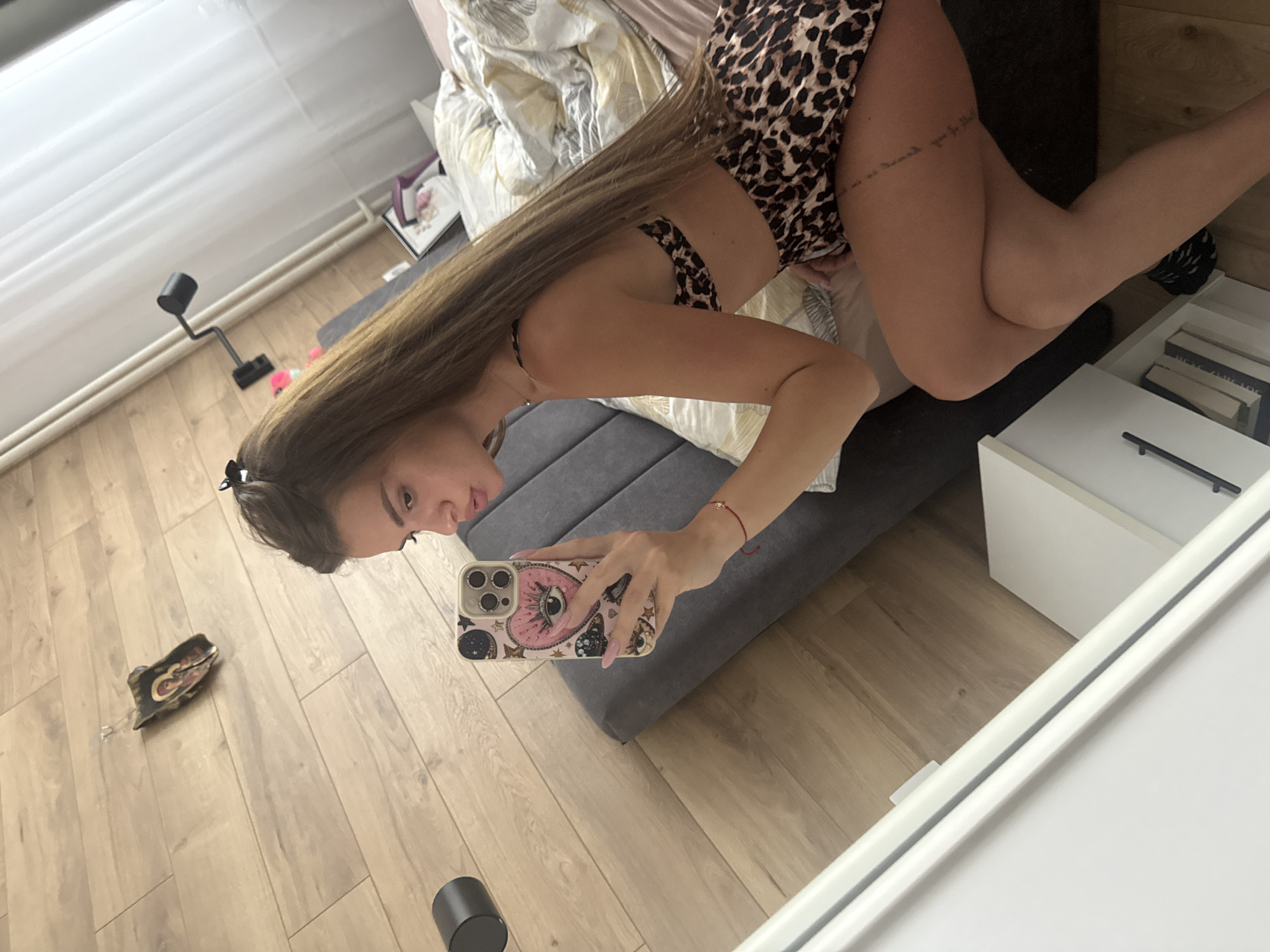 A selfie taken in an indoor setting shows a woman in a leopard-print bikini. She is lying on a bed with a gray headboard, taking the photo with a phone with a pink case. The bed is partially covered with patterned bedding. The room has light-colored wooden floors. There is a bedside table with a drawer next to the bed, as well as a black object on the floor. The location is Unknown, Unknown.