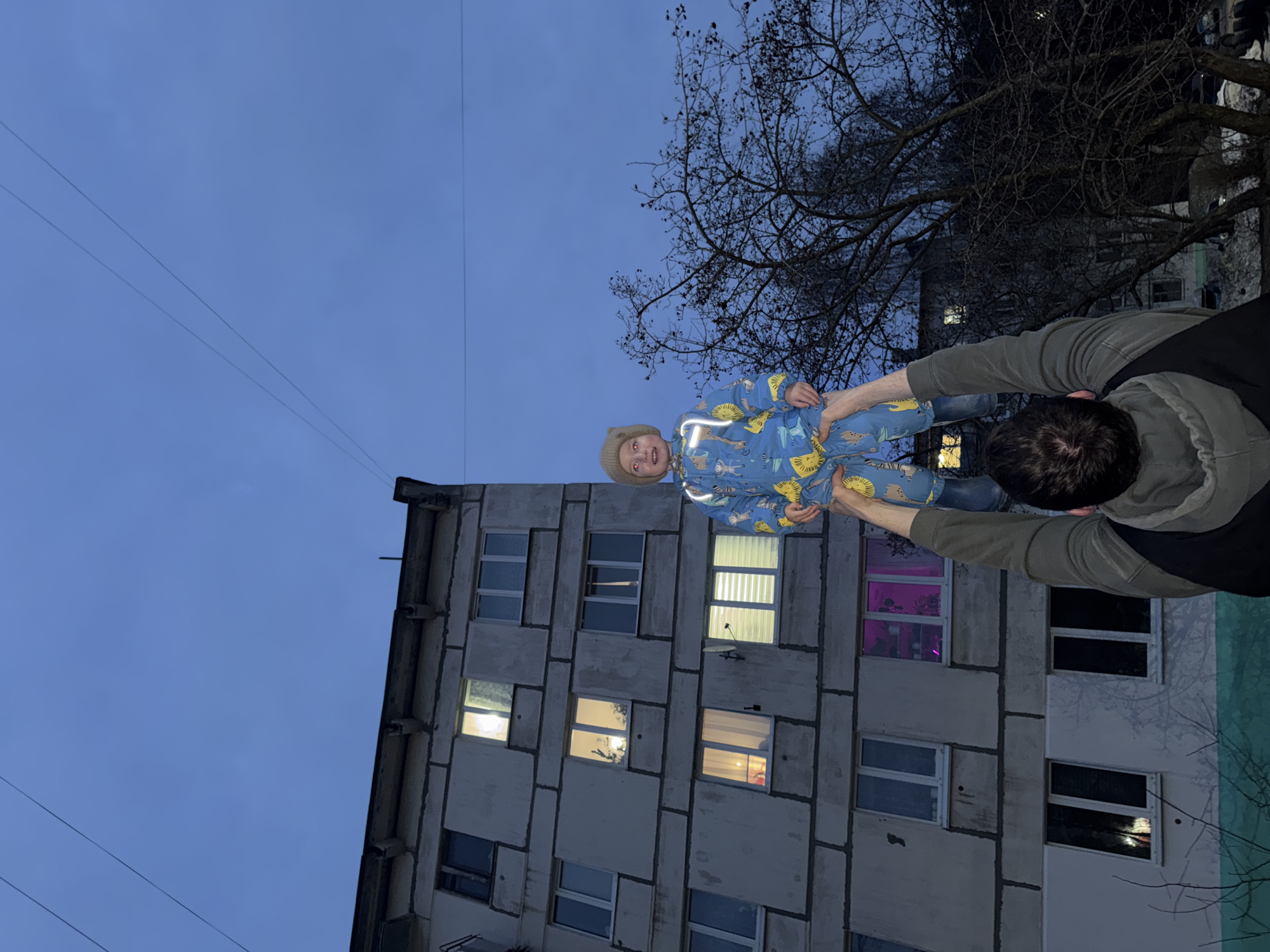 A man in a green jacket and black vest is holding a child in a blue animal-print jumpsuit aloft in front of a grey, Soviet-era apartment building at dusk. The child is smiling. The building has several windows, some lit and one with pink lighting. Bare tree branches are visible on the right. Overhead power lines are in the sky. The location is likely Chaikovsky, Russia, based on the context.