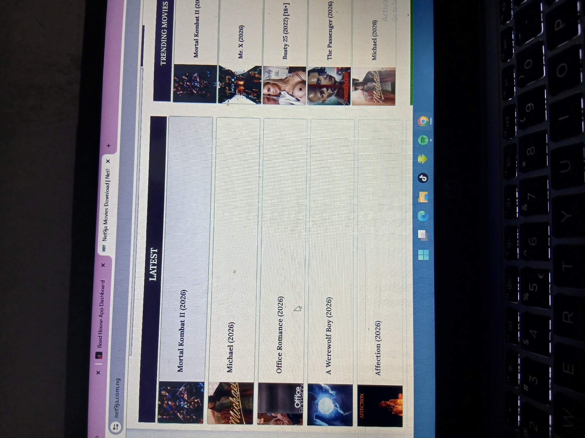 The image shows a laptop screen displaying a website with a list of movies. The website appears to be related to movie downloads, with categories like "LATEST" and "TRENDING MOVIES." Several movie titles are visible, including "Mortal Kombat II (2026)," "Michael (2026)," "Office Romance (2026)," "A Werewolf Boy (2026)," and "Affection (2026)." The laptop is placed on a dark surface, likely a keyboard, as indicated by the visible keys. The scene does not provide any information about the location in the city of Howayi, Nigeria.