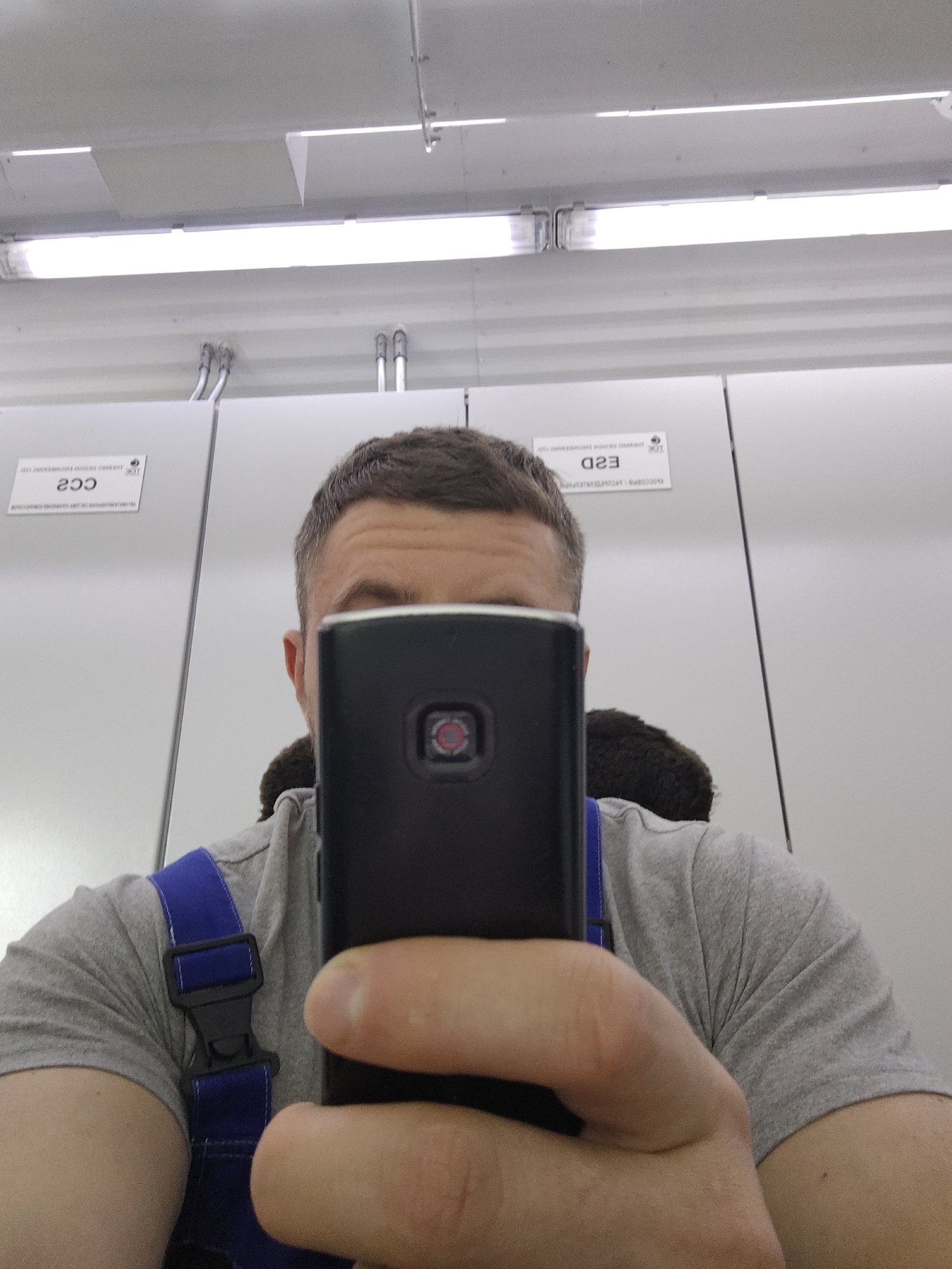 A person takes a close-up selfie with a black smartphone, obscuring their face. They are wearing a grey t-shirt and blue overalls. Behind the person are plain white cabinets with small informational labels on them and fluorescent lights on the ceiling. The location is indoor.