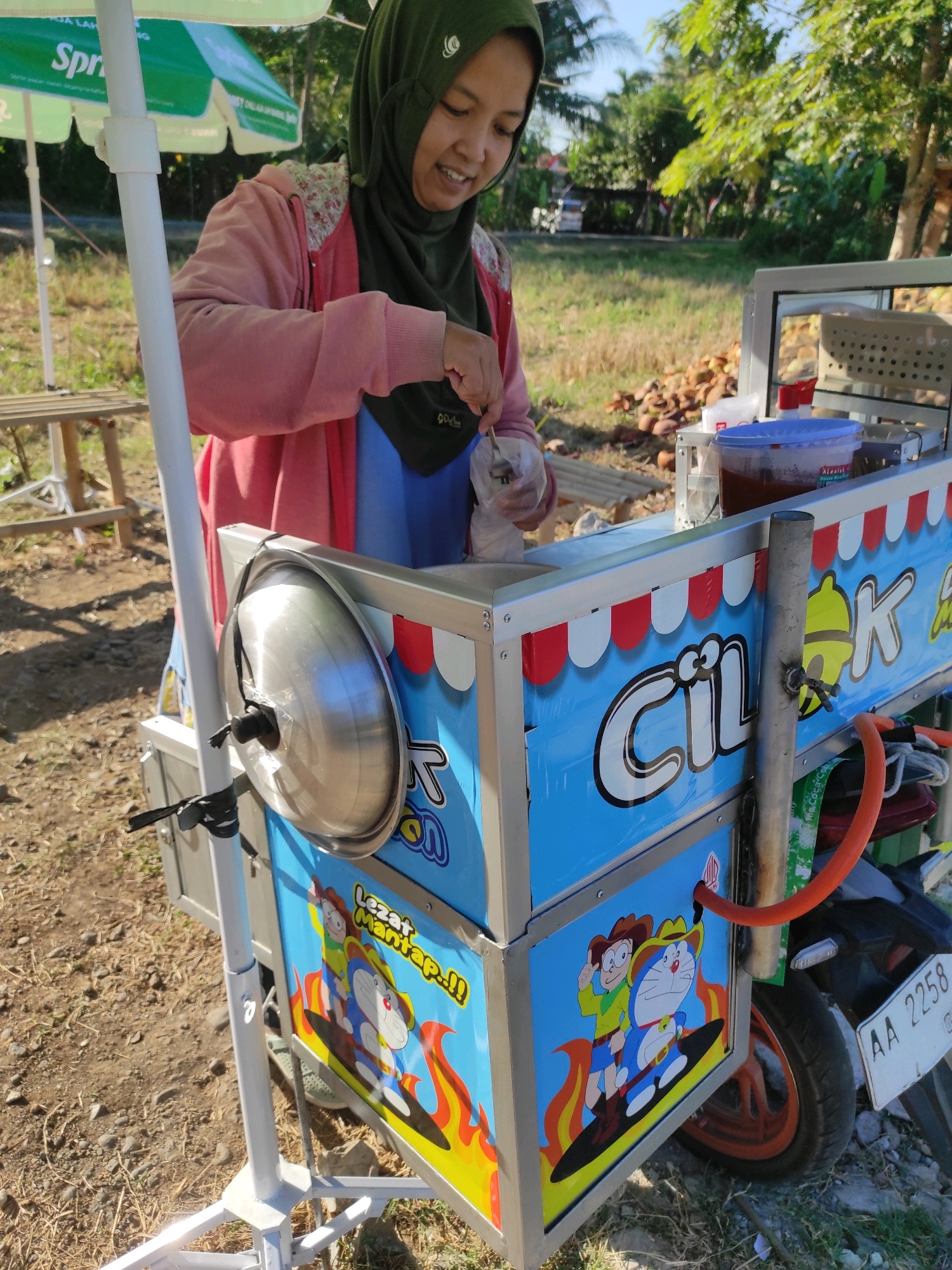 A young Indonesian woman wearing a green hijab and a pink jacket is operating a food cart in Purwodadi, Indonesia. The cart is decorated with blue panels featuring cartoon characters, including Doraemon, and the text "Cilok". A gas cylinder is attached to the cart. The background shows a rural setting with trees and dry vegetation. The woman is holding a bag and a utensil, seemingly preparing food for a customer. The scene is illuminated by natural sunlight.