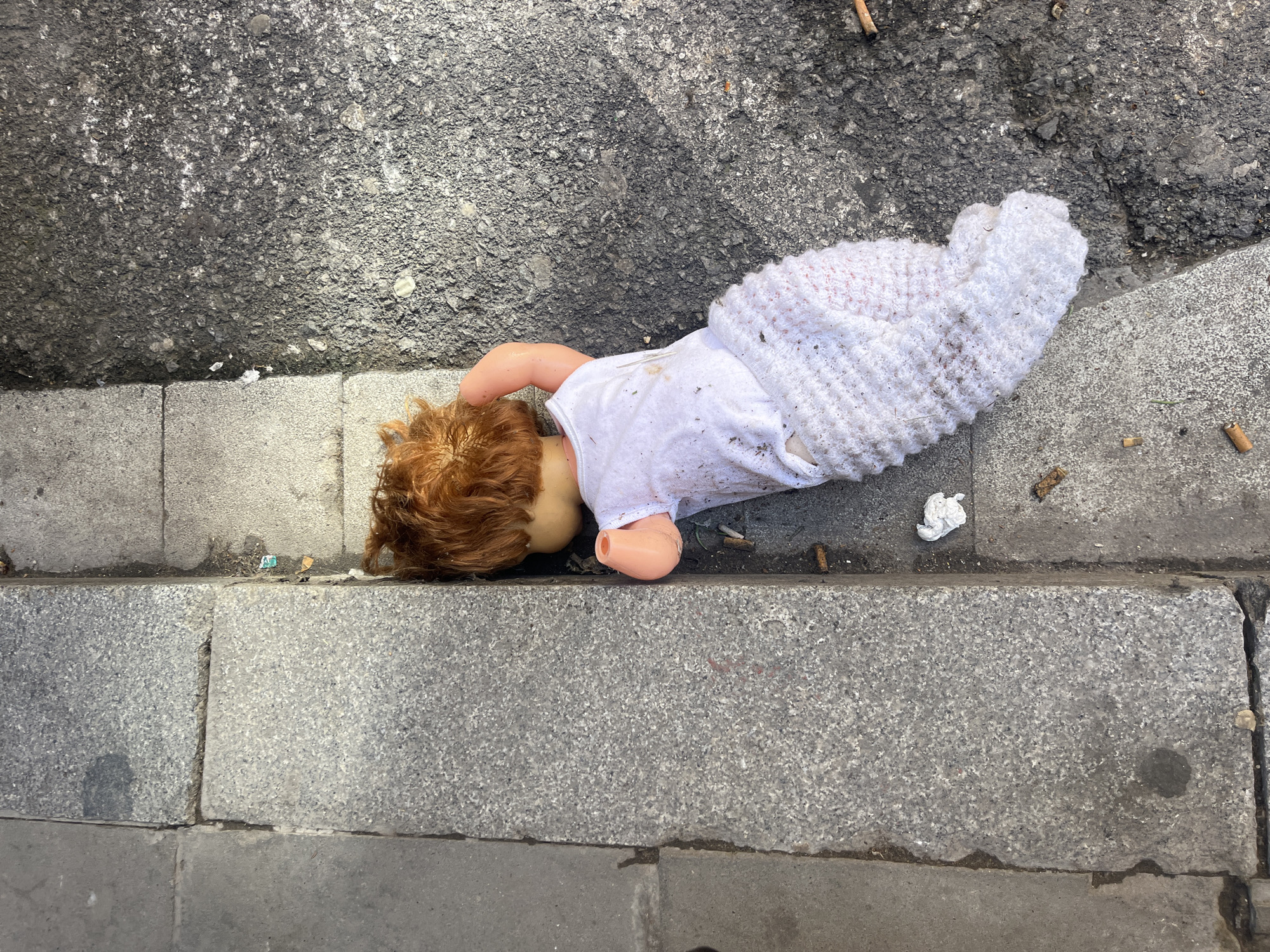 A discarded doll lies on its side on an urban street, partially on dark gray asphalt and partially on a light gray granite curb. The doll has reddish-orange hair and is dressed in a white top, with its lower body wrapped in a white knitted blanket. Its head faces the asphalt, and one arm is bent at the elbow. The doll appears somewhat dirty.
The immediate surroundings feature several cigarette butts scattered on the asphalt and along the curb, alongside a crumpled white tissue and other small bits of debris. The lighting indicates it is daytime. No specific activity is taking place; the image captures a static scene of an abandoned item. The location context for this image is Barcelona, Spain. There is no visible text in the image.