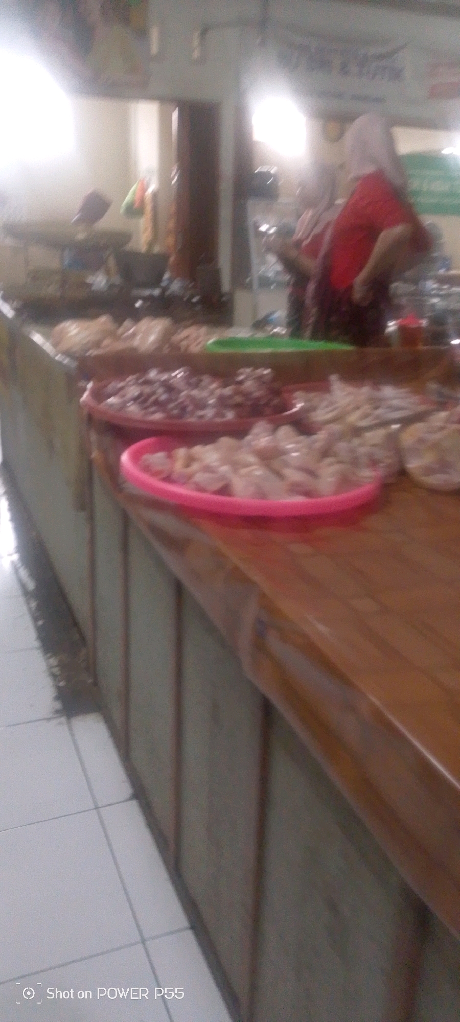 The image depicts a market stall with an assortment of raw meats displayed in plastic containers. In the foreground, a pink plastic container holds chopped raw chicken pieces. Behind this, a red plastic container contains what appears to be raw minced meat or small pieces of meat. Further back, other meats are visible, along with a green plastic tray holding some produce. Two women, one wearing a red headscarf and red top, and another partially visible in a light-colored headscarf and dark top, are positioned behind the counter. The stall has a wooden countertop and the background shows a blurred market environment with lighting. The floor is tiled. The image contains a watermark "Shot on POWER P55" in the bottom left corner.