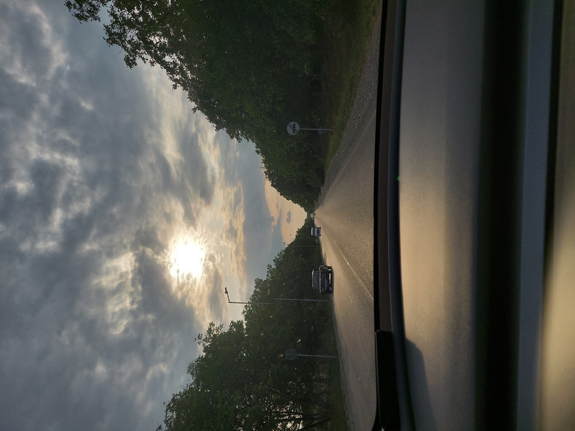 The image, taken from the interior of a vehicle, shows a road lined with dense green trees on both sides. The sun is visible through a cloudy sky, casting a warm glow on the scene. Two cars are traveling on the road, one in the foreground and another further down. There are also streetlights and traffic signs visible along the road. The foreground of the image is dominated by the interior of the car, including the dashboard. The scene depicts a peaceful drive through a natural landscape with a hint of urban infrastructure. Based on the provided metadata, this scene is located in Mirikənd, Azerbaijan.
