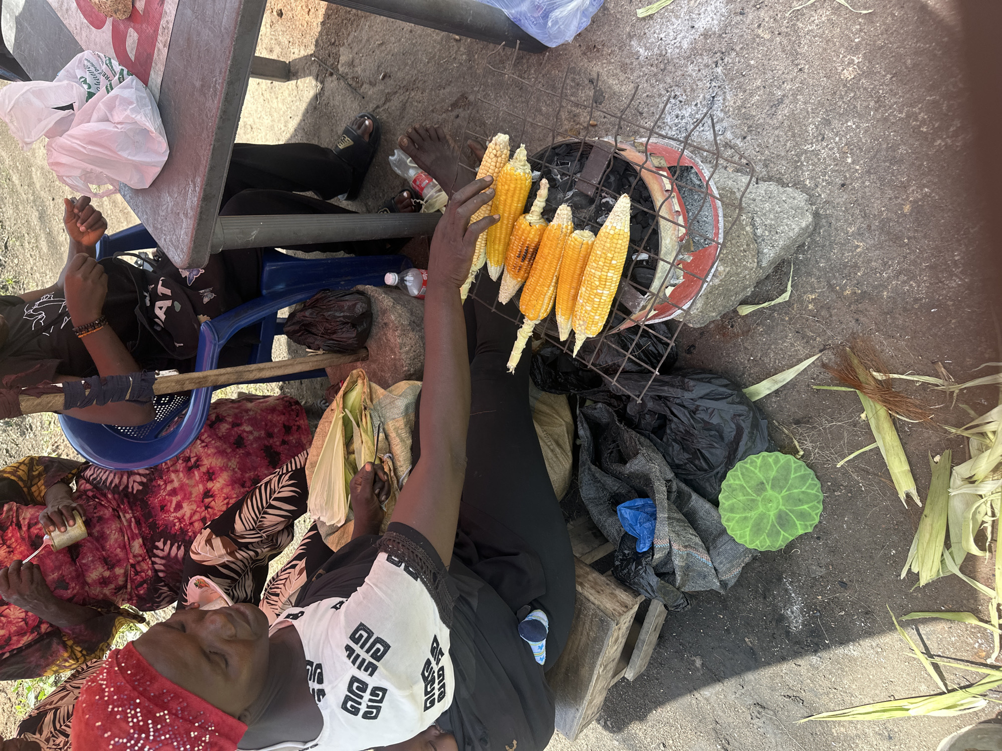 In Gura Top, Nigeria, a woman with a red headscarf is seated outdoors, preparing to roast corn over a small charcoal grill. She is wearing a white shirt with a black graphic print and black pants. Her hands are positioned near several ears of corn that are already on the grill. To her left, more ears of corn are wrapped in their husks, alongside some other food items. The ground is a mix of dirt and concrete, with various discarded materials visible, including plastic bags, a green decorative item, and corn husks. In the background, other individuals are present, and a blue plastic chair is visible. A metal table with white plastic bags on it is also in the scene. The overall setting appears to be a casual outdoor market or street food stall.