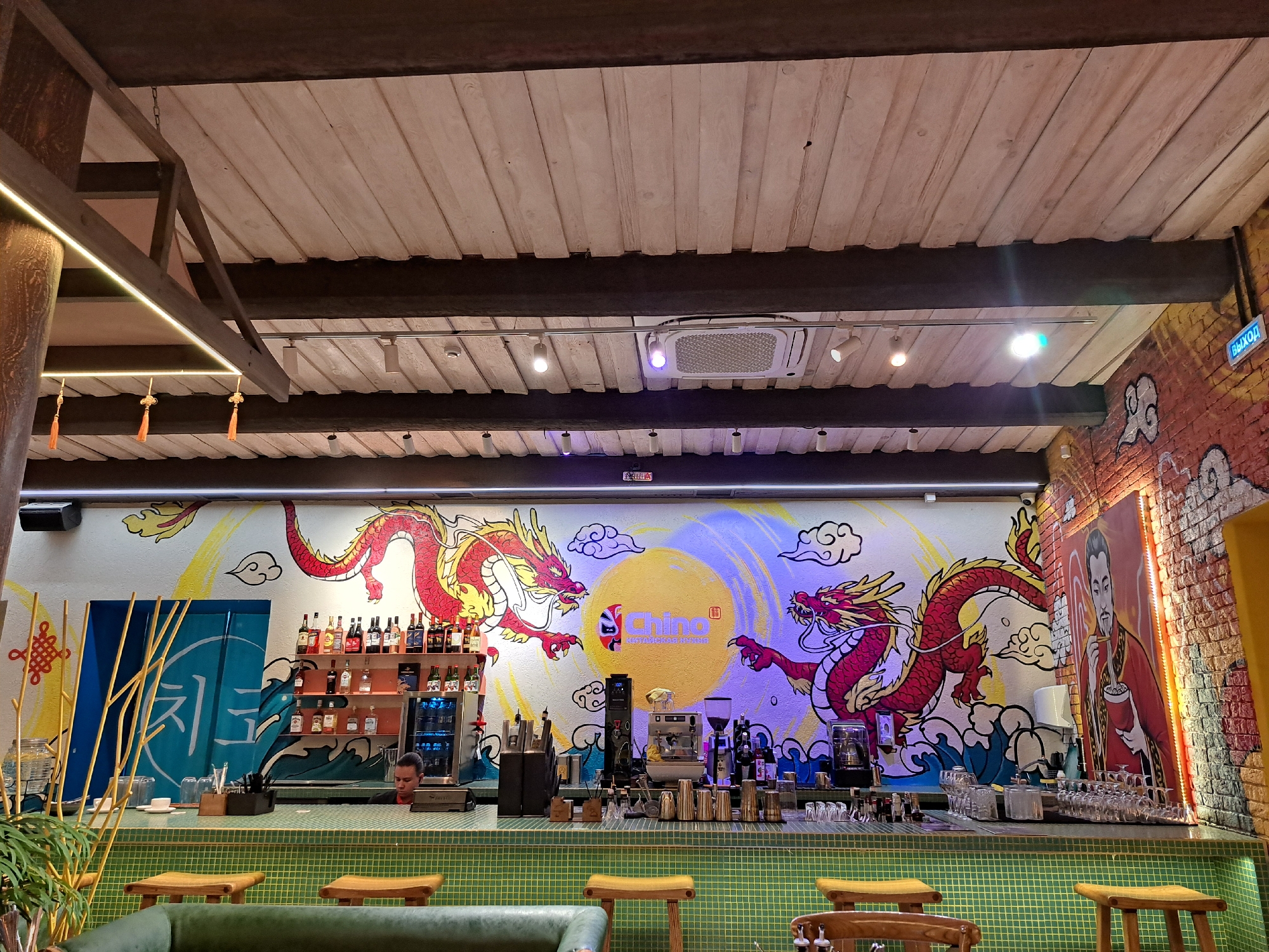 The image depicts the interior of the "Chino" restaurant located at Karla Libknekhta Street, 7, Irkutsk, Russia. The scene is a bar area with a prominent mural on the wall featuring two red dragons, a yellow sun, and clouds. In the foreground, a green tiled bar counter is visible with several wooden stools. Behind the counter, a selection of alcoholic beverages is displayed on shelves, and bar equipment such as a coffee machine and blenders are present. A male bartender is seen working behind the counter. The ceiling is constructed with wooden beams and planks, and an air conditioning unit is installed. A brick wall with graffiti and a painting of a man in traditional attire is on the right side of the image. An illuminated "Exit" sign is also visible.