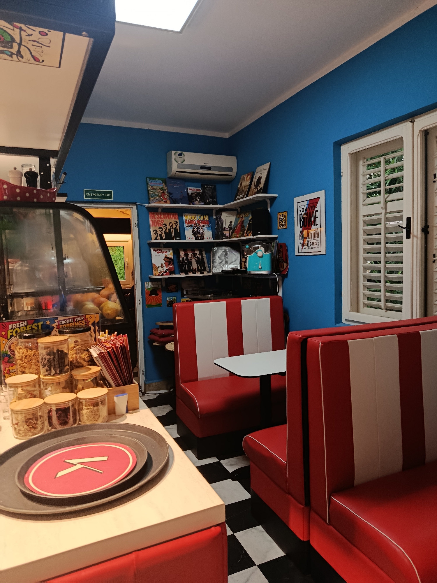 This image depicts the vibrant interior of a retro-themed diner or cafe, characterized by its bright blue walls, red and white striped booth seating, and a classic black and white checkered floor. The space appears empty, suggesting it is either closed or awaiting customers.
On the left, a counter features a display case filled with fresh produce like oranges and lemons. Below, several glass jars contain various dry toppings, labeled "FRESH FOREST" and "CHOOSE YOUR TOPPINGS." A box of red and white striped paper sleeves or straws is also present. In the foreground on the counter, two dark plates are stacked, with the top one holding a red circular placemat featuring a white stylized "K" or similar symbol.
The blue back wall is adorned with white shelves holding books, records, and posters. Visible titles include "RESERVOIR DOGS," "MAKU CHAO RADIO REMB," "DILAN DOG," and "NOE." A light blue appliance, possibly a popcorn machine, sits on a shelf, and an air conditioning unit is mounted above. A prominent framed poster advertises "DAVID BOWIE" with dates "JULY 3-1973" and other text like "KINGSTON DUST" and "KETS (1.59/92.00." A small framed image of a bull is also on the wall.
To the right, a large window with white louvered shutters is partially open, revealing green foliage outside, indicating it is daytime. An "EMERGENCY EXIT" sign is visible above a doorway on the left side of the blue wall. The red and white booths, arranged with small white tables, evoke a nostalgic 1950s American diner aesthetic. The overall scene suggests a cheerful and inviting atmosphere for a cafe or diner in Herceg Novi, Montenegro.