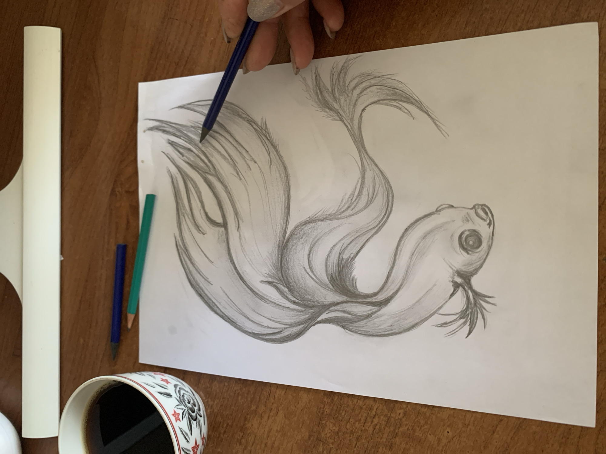 A close-up, overhead shot shows a hand with glittery fingernails holding a dark blue pencil and drawing a sketch of a fish on a white piece of paper. The fish is stylized, with flowing fins that fill most of the paper. The drawing is done in graphite pencil. To the left of the paper, two colored pencils, one green and one blue, lie on a wooden surface. A white object, possibly a clipboard, is also on the left side of the frame. In the bottom left corner, a white ceramic mug with a black and red floral pattern is filled with dark liquid, likely coffee. The scene appears to be set indoors on a wooden table. The lighting suggests it could be daytime. There is no visible text in the image.