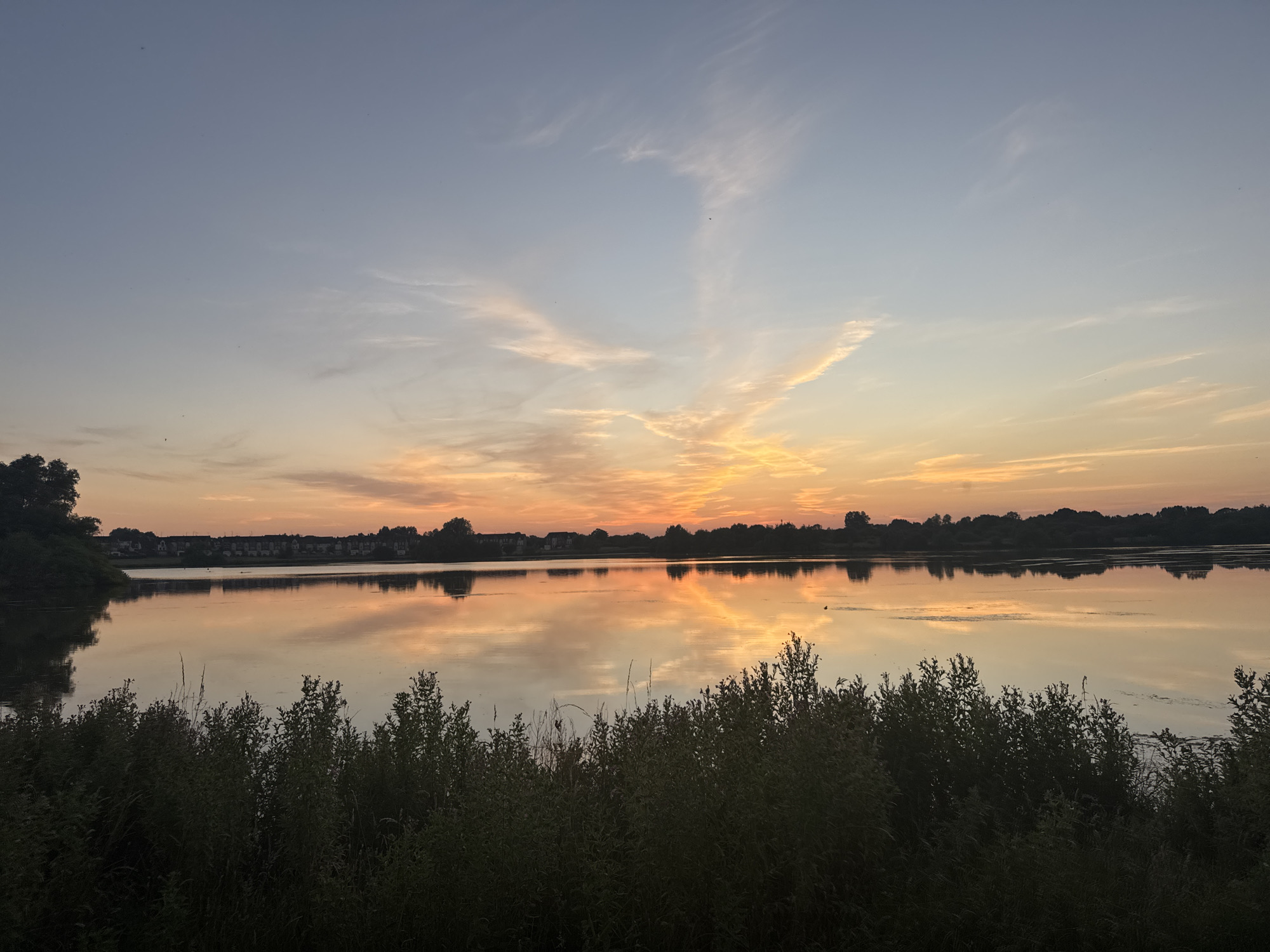 This is a photograph taken at sunset of a lake in Craigavon, United Kingdom. The sky is a gradient of colors, transitioning from a pale blue at the top to warm oranges and yellows near the horizon. Wispy clouds are scattered across the sky, catching the light of the setting sun. The lake's surface is calm and reflects the colors of the sky, creating a mirror image of the sunset. In the distance, a line of trees and houses can be seen along the far shore of the lake. In the foreground, dense green foliage and bushes line the edge of the lake. The overall impression is one of tranquility and natural beauty.