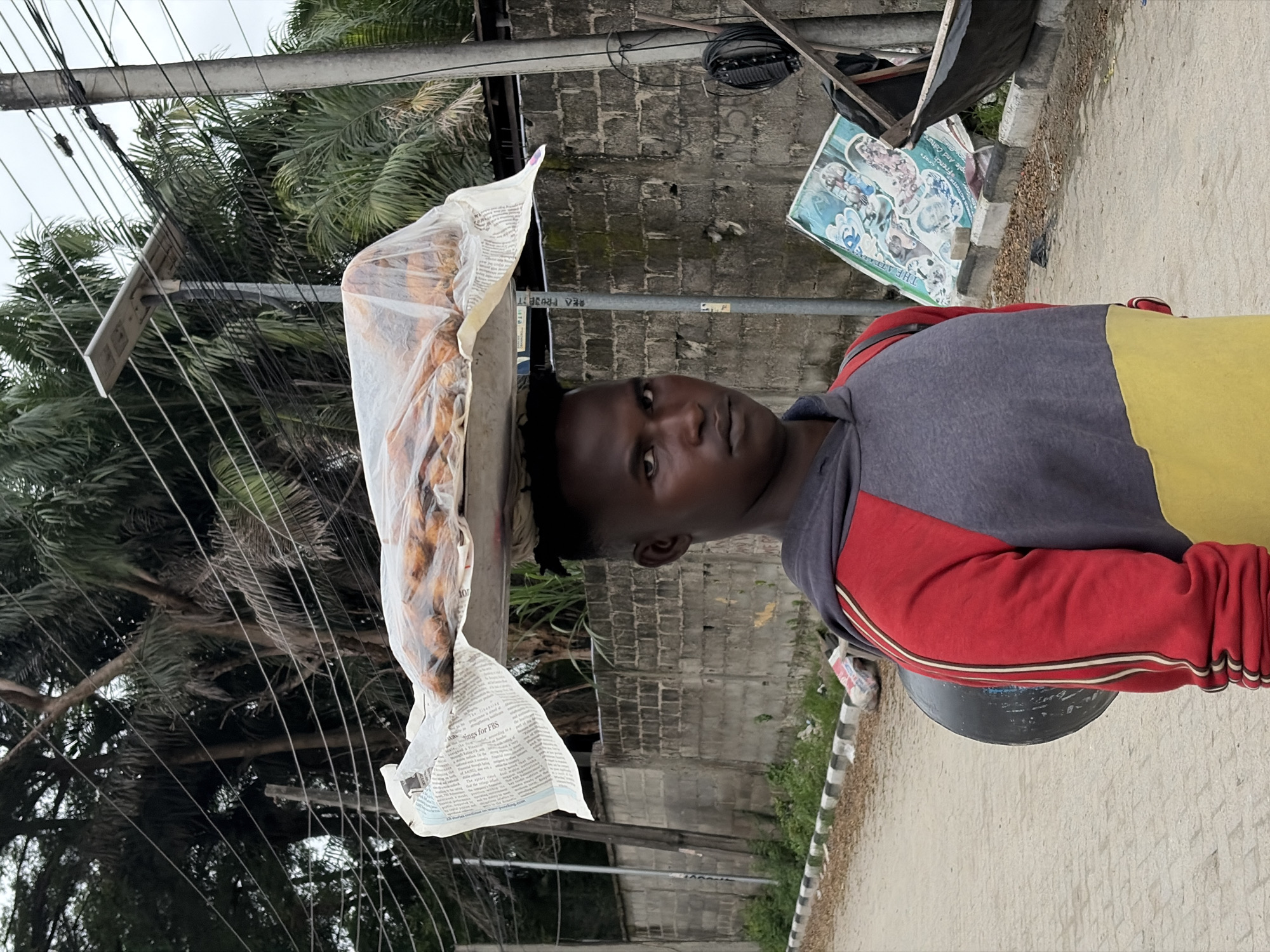 A young man stands on a paved street in Igboefon, Nigeria, with a tray of food balanced on his head, covered in plastic wrap and newspaper. He wears a red and grey hooded sweatshirt with a yellow lower portion and a black circular object slung over his left shoulder. Behind him, a grey brick wall runs along the street, with lush green palm trees and other vegetation visible above it. A utility pole with electrical wires and a solar panel is positioned to the right of the man. In the background, to the right of the wall, a large, colorful poster is propped up, and a dark, open-topped structure with a black tarp is also present. The ground is a light-colored paving material, with a concrete curb and a strip of grass and dirt bordering the street. The sky appears overcast.