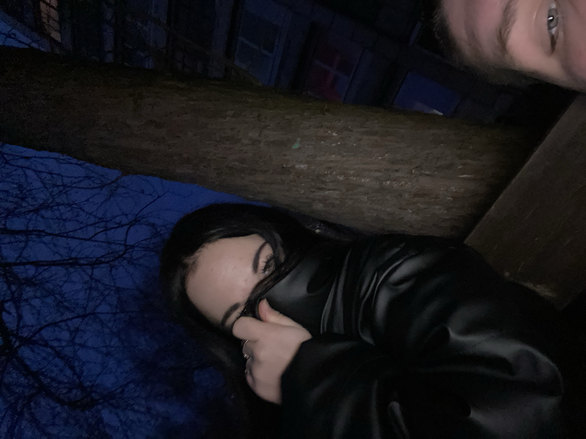 A low-angle, close-up photograph taken at night depicts two individuals. One person, a young woman, is in the foreground wearing a black jacket and partially covering her face with her hand. Rings are visible on her fingers. Her dark hair frames her face, and her eyes are visible. Another individual, partially visible at the top right corner, shows a small part of their face including an eye. The background is a mix of dark, unclear objects. In the upper section of the background, indistinct building structures can be seen, including what appears to be a window with a red frame. To the left, bare tree branches are silhouetted against the night sky. The location is identified as Chaikovsky, Russia. The lighting is dim.