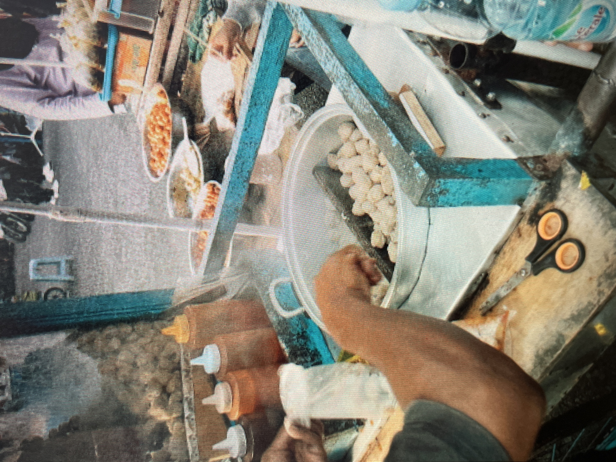 In Sejahtera, Indonesia, a street food vendor is preparing food at a stall. The vendor, partially visible with an arm reaching into a large pot, is likely serving customers from a busy street. The stall is equipped with various containers of ingredients and condiments, including two squeeze bottles of sauce. Nearby, a pair of scissors rests on a wooden surface. The scene depicts a common urban marketplace setting, bustling with activity and the aroma of local delicacies.