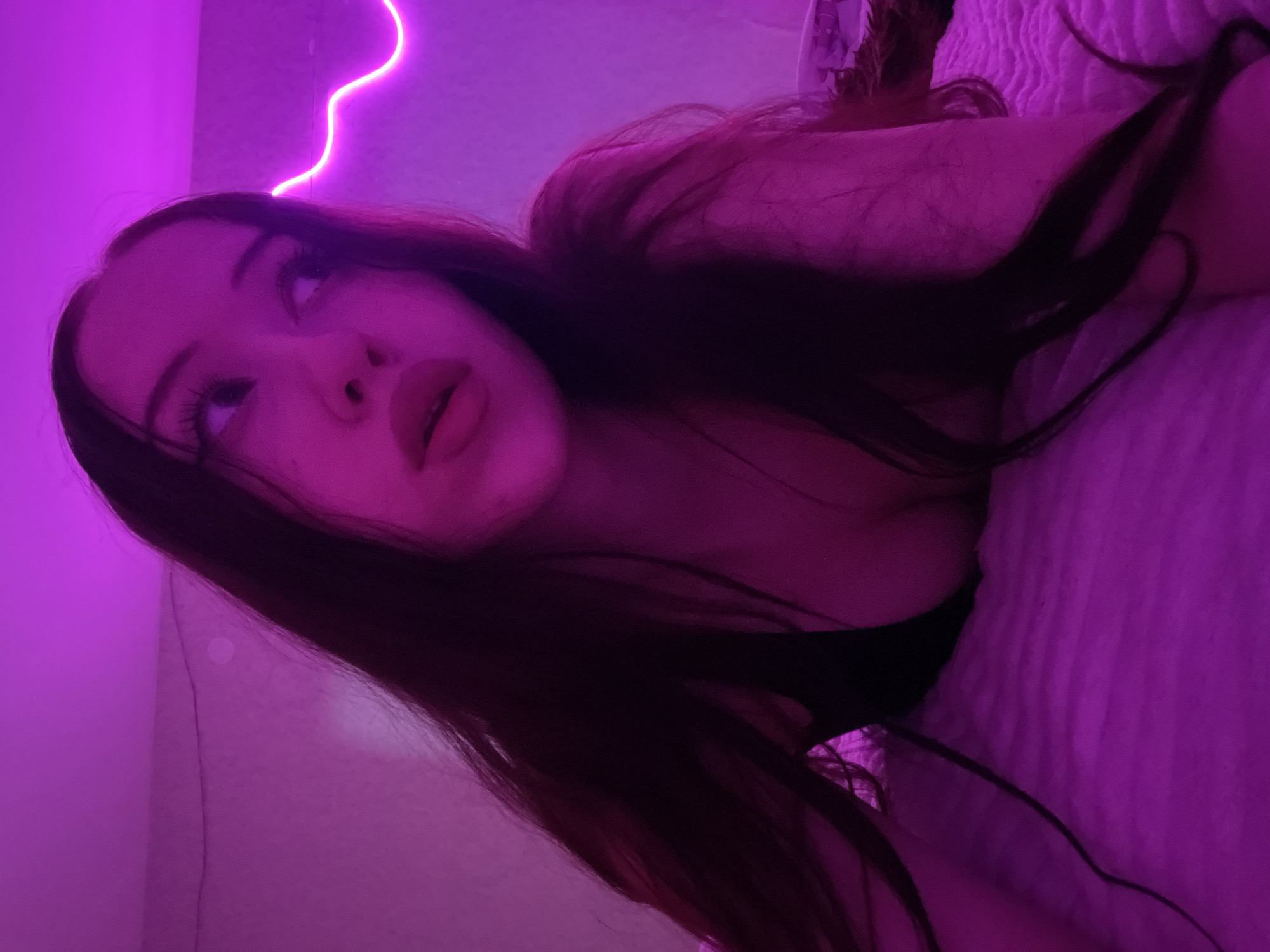 The image shows a young woman with long dark hair lying on a bed in a room illuminated by pink neon lighting. She is wearing a dark-colored top that reveals her cleavage. Her head is turned to her right, and she is looking upwards and to the side. The background is a wall with a wavy neon light strip. The bedspread appears to be textured and is also illuminated by the pink light. There are no visible details that would indicate the location in Votkinsk, Russia.