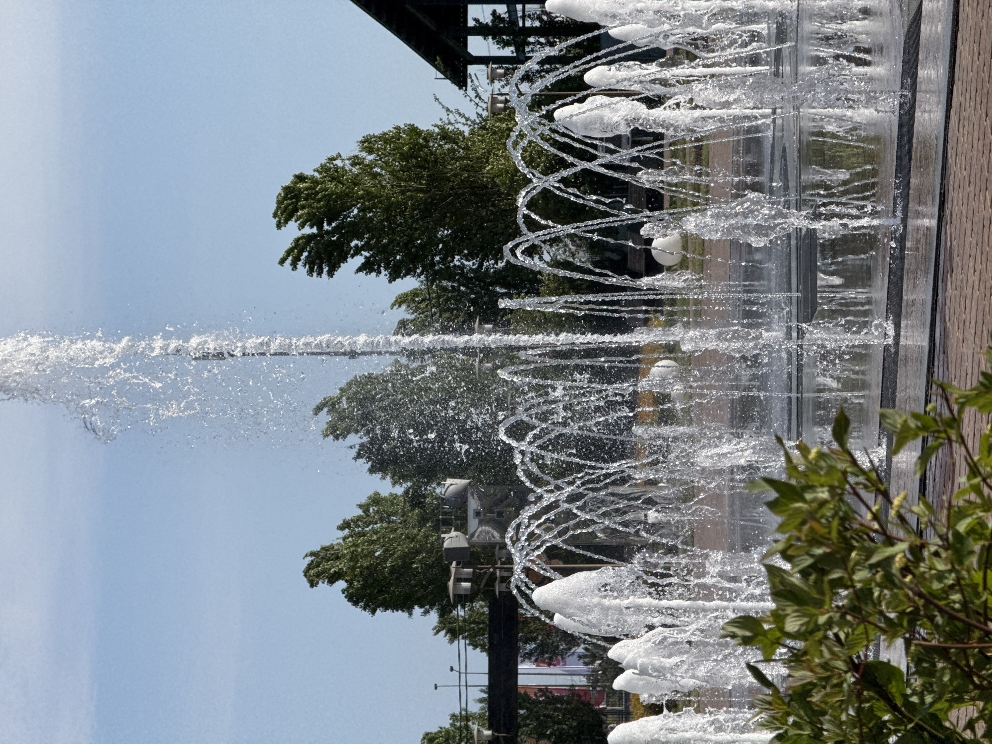 The image shows a fountain in Rostov-on-Don, Russia. The fountain features multiple streams of water shooting upwards and curving, with some forming spiral shapes. A tall, concentrated water jet shoots upwards on the left side of the frame. Behind the fountain are trees and infrastructure elements. To the right, there's vegetation obscuring part of the fountain's edge. The sky is pale blue and the composition focuses on the water features and the urban background.