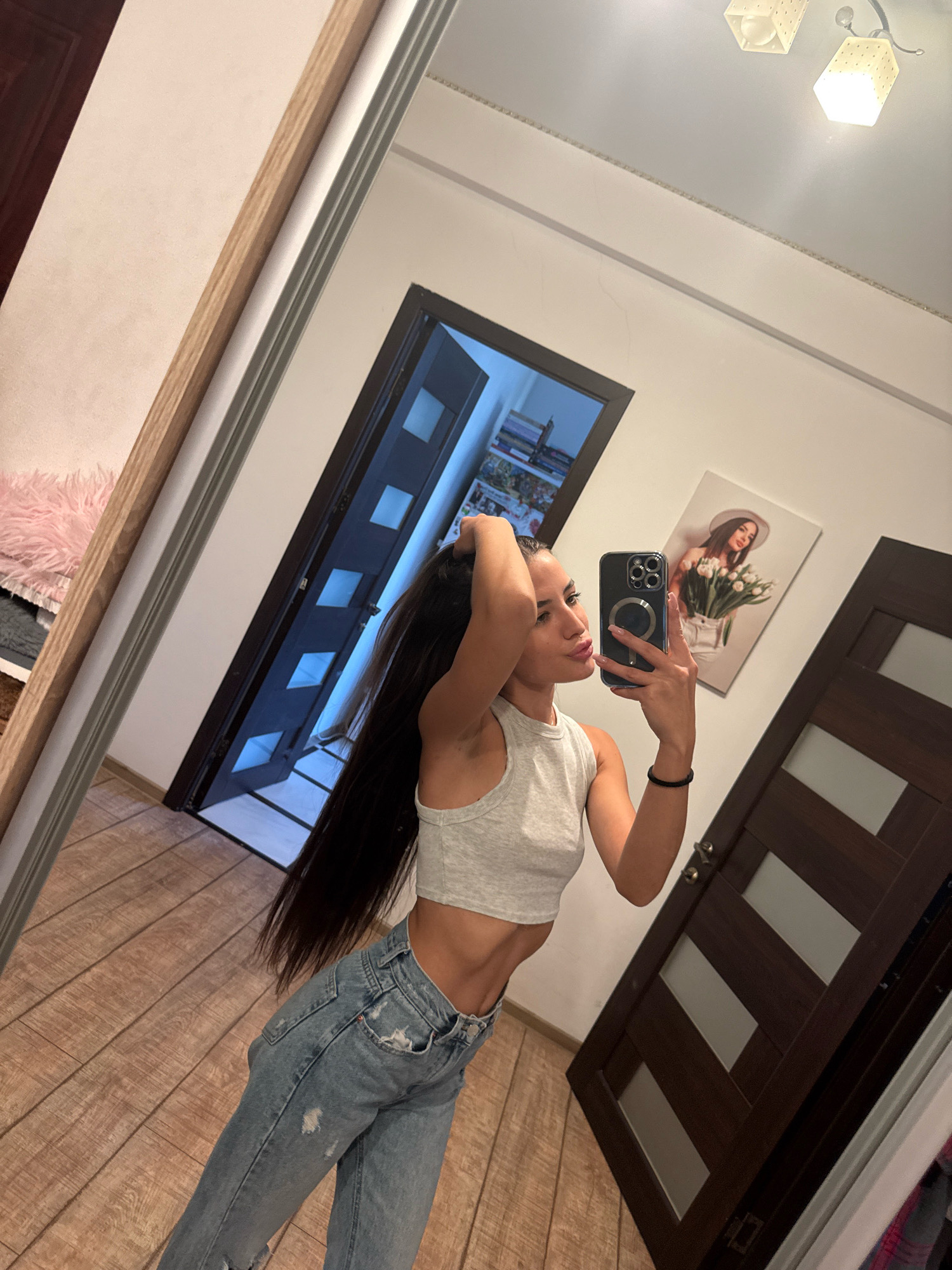 A young woman with long, dark hair pulled back is taking a mirror selfie in what appears to be a residential hallway or room. She is wearing a grey crop top and ripped blue jeans. She is holding her phone, a black iPhone with a silver camera ring, in her right hand, and her left hand is in her hair. In the reflection of the mirror, a door with diamond-shaped glass panes is visible, along with a framed picture of a woman holding tulips on the wall. The floor is made of light brown wood planks. The lighting suggests it is daytime, with a ceiling light fixture visible. The overall impression is a casual, indoor setting. This scene is not identifiable as being in Chișinău, Moldova, or any specific city.