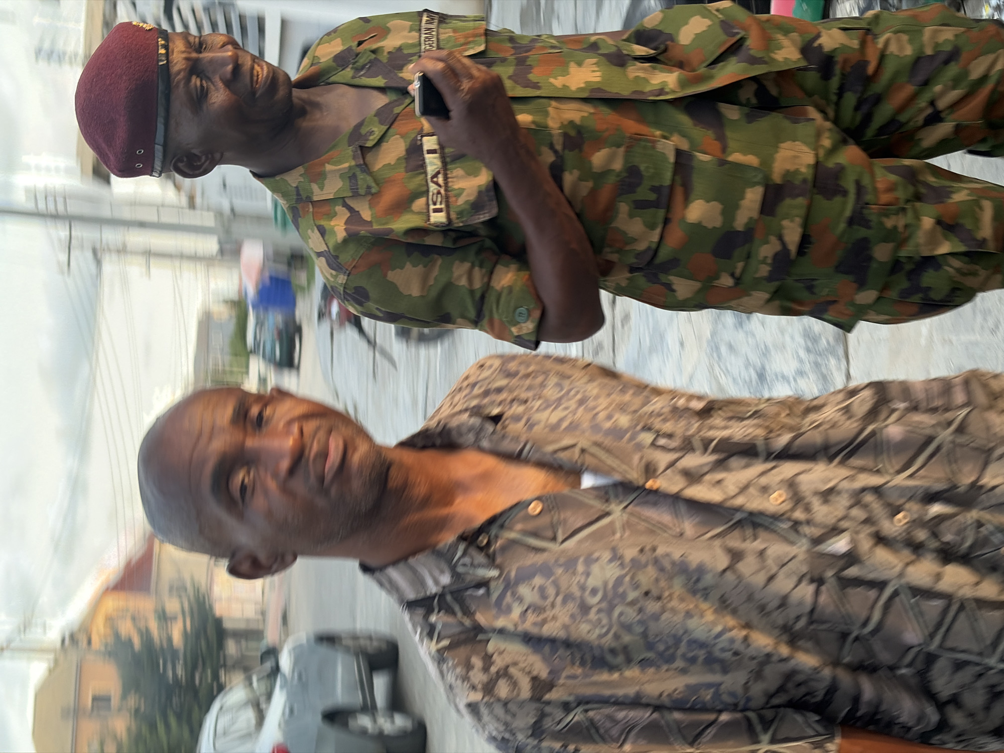 The image shows two African men, likely Nigerian, with a blurred urban background suggestive of the city of Igboefon, Nigeria. One man is wearing a Nigerian military uniform, with a maroon beret and camouflage pattern, holding a device. The name "ISA" is visible on his uniform. The other man is wearing a patterned shirt. Cars and buildings are blurred in the background.