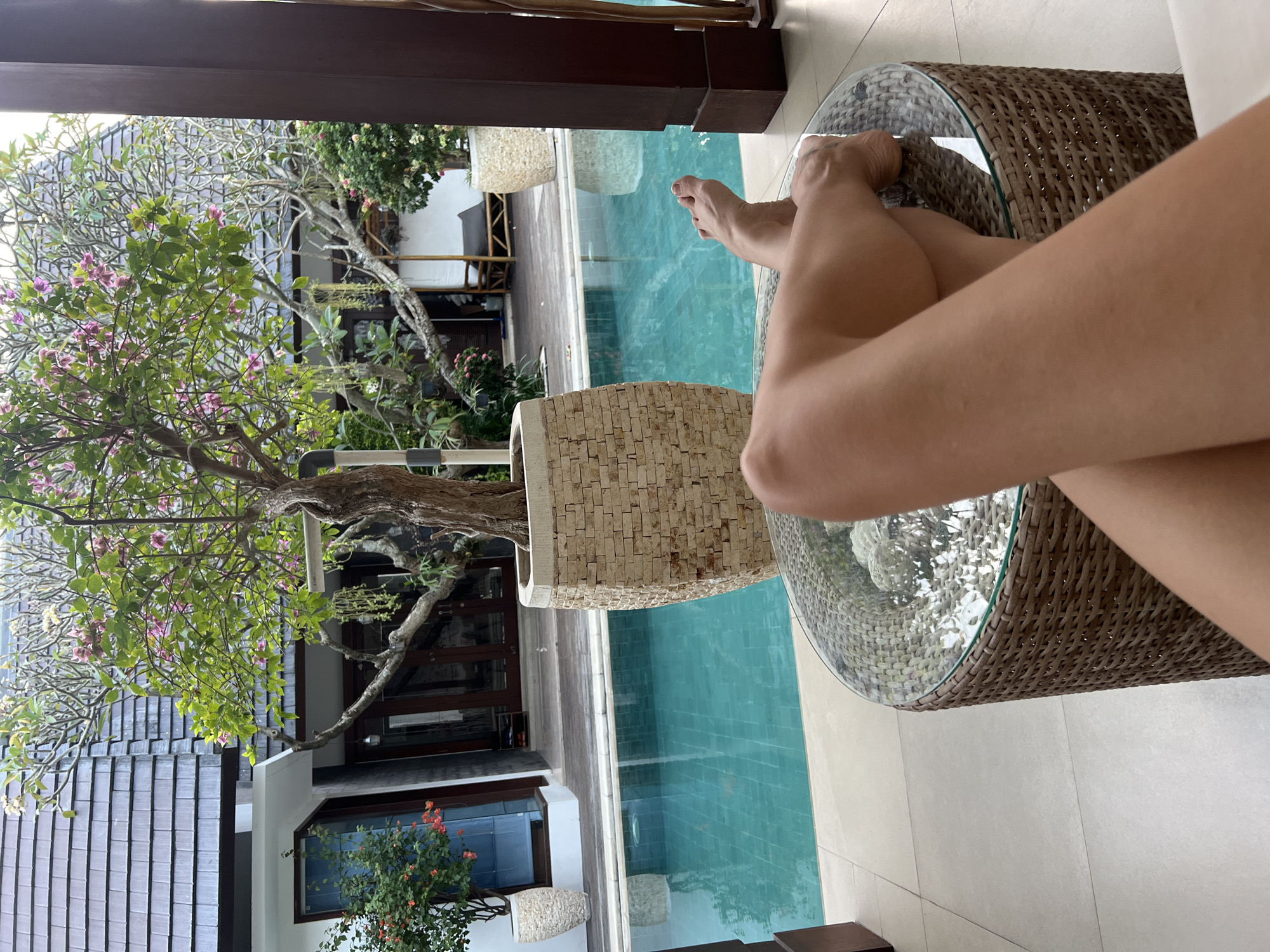Captured in North Kuta, Indonesia, the image features crossed legs resting on a round glass-topped rattan table. A tiled swimming pool is visible, flanked by a textured planter. A tree with purple blossoms grows nearby, in the background, a building with dark roof tiles and window frames is visible.