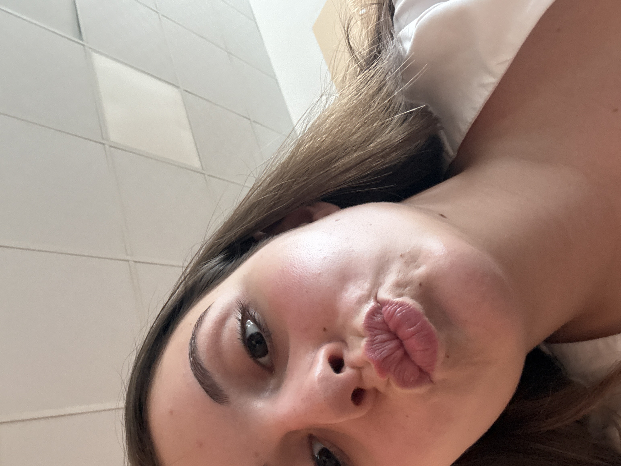 A close-up selfie captures a young woman's face and upper torso from a low, upward-looking angle. Her head is tilted, with her dark brown hair falling to the left side of the frame. She has distinct, dark eyebrows and dark eyes, one looking directly at the camera, the other partially obscured by the angle. Her lips are pursed into a pronounced "kiss face" expression, appearing plump and well-defined. Her skin shows a clear complexion with a subtle flush on her cheeks. She is wearing a white garment, visible on her right shoulder and neck. The background consists of a light-colored, grid-patterned surface, possibly tiled or paneled, indicative of an indoor setting like a wall or ceiling. The image conveys a playful and informal mood. No text is visible. The scene is located in Burgas, Bulgaria.