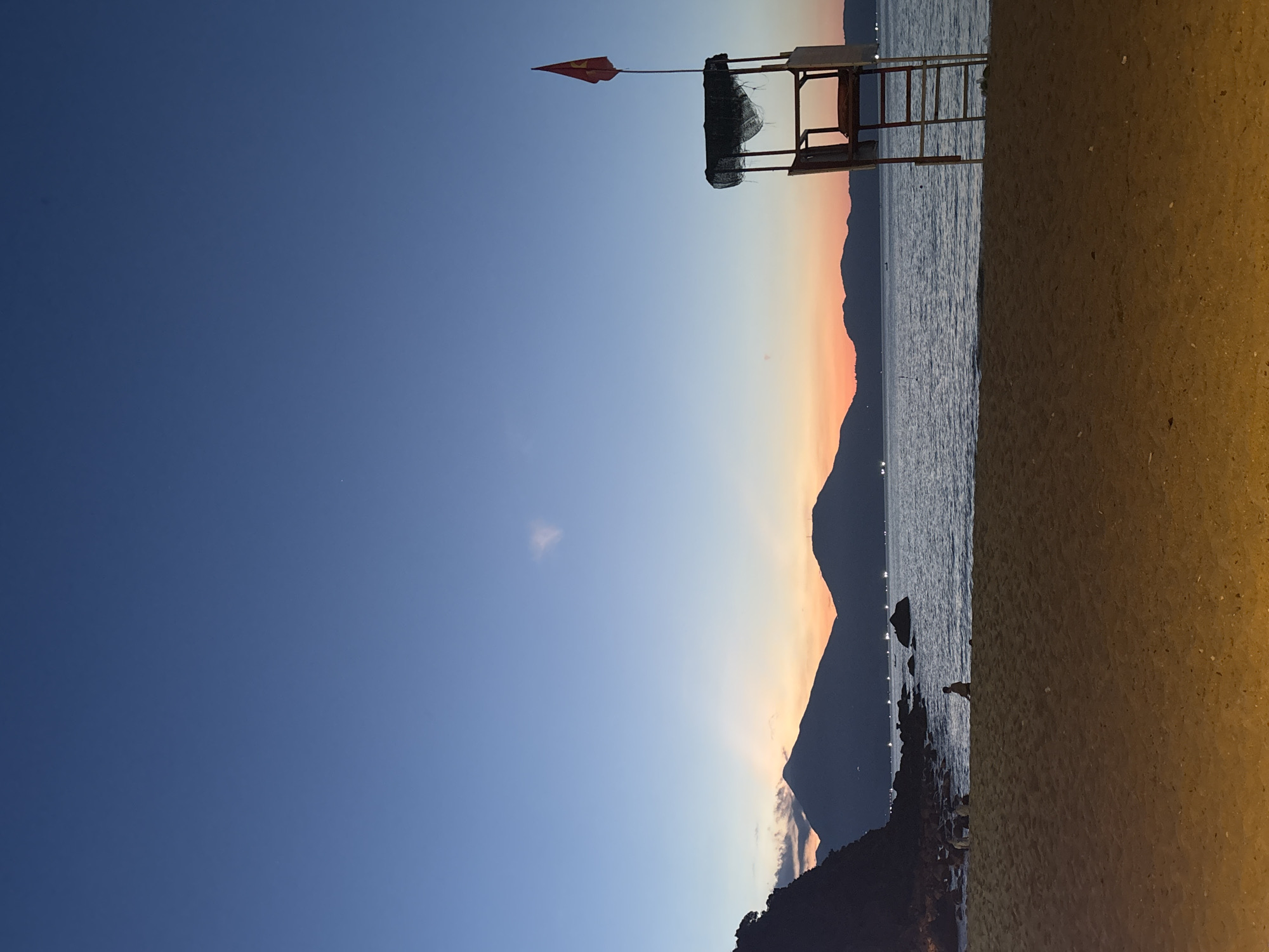 The image depicts a beach scene, likely at dusk. A lifeguard stand with a red flag is visible in the foreground. The backdrop consists of a mountain range silhouetted against a sky transitioning from blue to orange hues. The water reflects light, suggesting a calm sea. The beach sand is a light color. The location is potentially in Đà Nẵng, Vietnam, considering the landscape characteristics. A bird stands on rocks at the shoreline.
