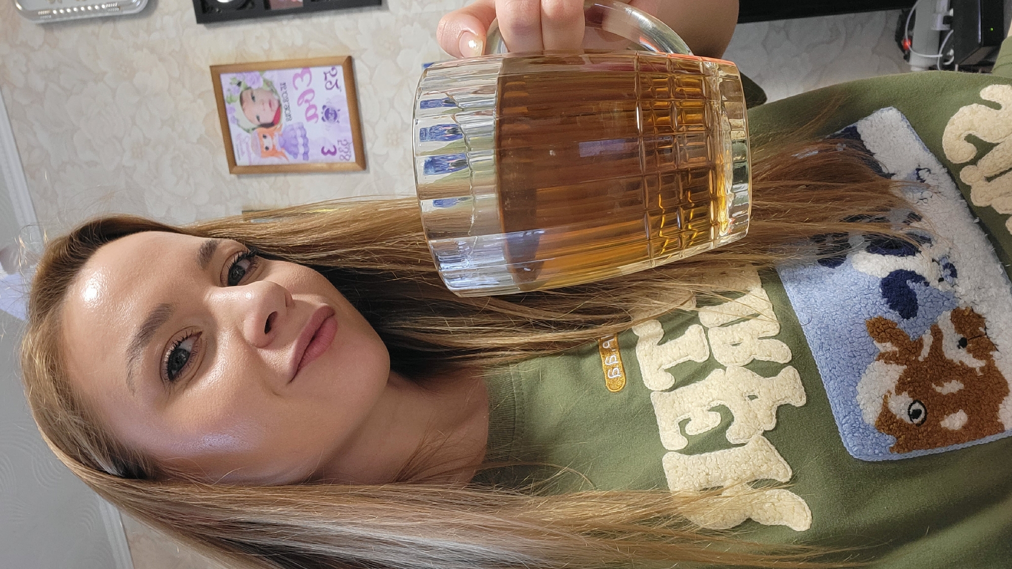 A woman with fair skin and light brown hair holds a glass mug filled with a light amber liquid, likely beer or iced tea, in front of a blurred background. She is wearing an olive green t-shirt with white stylized lettering and a patch featuring a cartoonish dog design in shades of blue, brown, and white. The background features a light-colored patterned wallpaper, a framed picture of a child, and an electronic device. The image was likely taken in Chaikovsky, Russia.