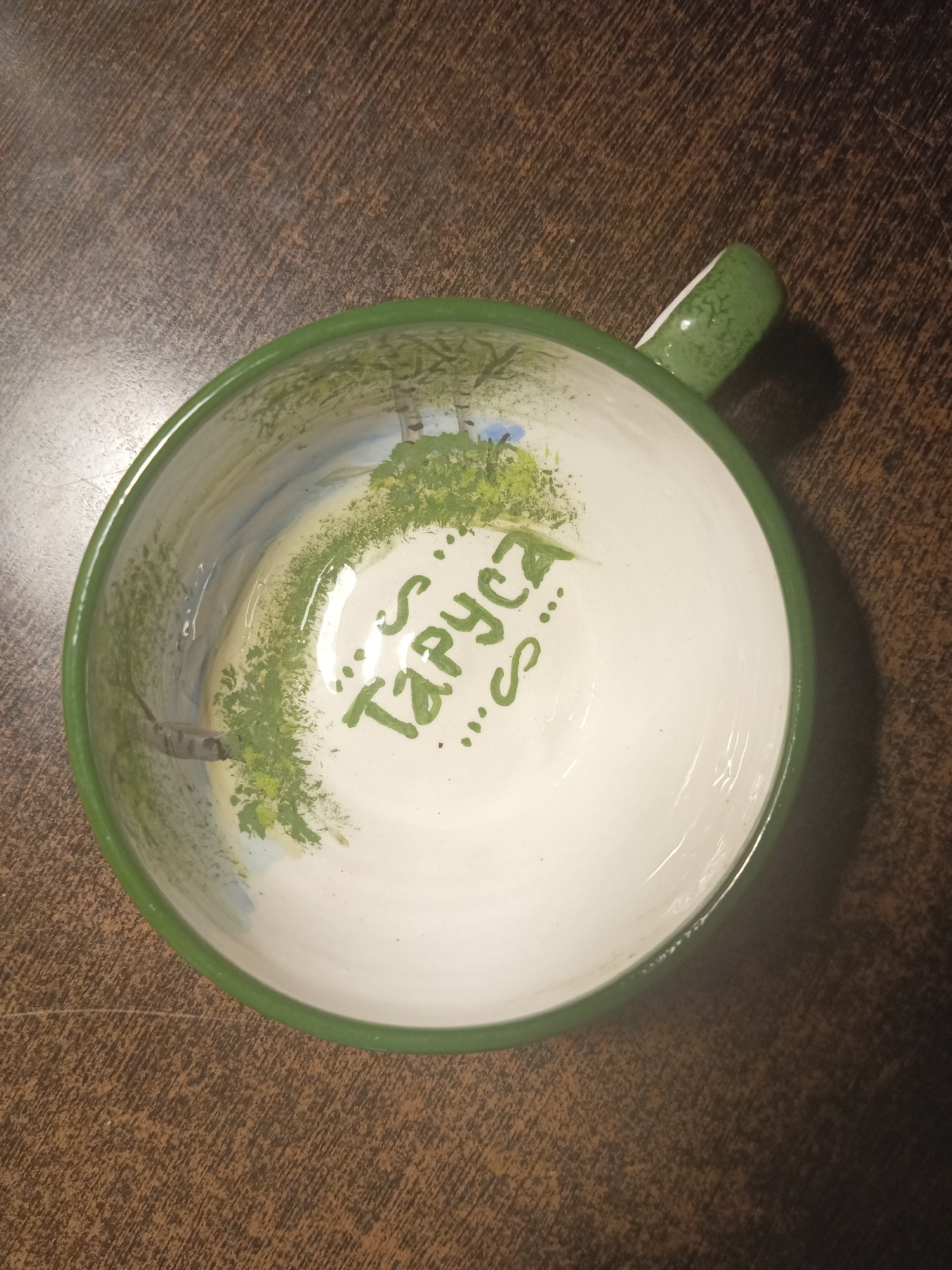 An overhead, close-up shot shows a white ceramic bowl with a green rim and handle. The interior of the bowl is painted with a landscape scene featuring trees and grass, and the words "S. Таруса" are written in green paint. The bowl sits on a dark brown, wood-grain textured surface. The image is dimly lit, with shadows visible. There are no people or animals present. The scene is indoors, likely on a table. The city of Obninsk, Russia is not depicted or referenced in the image.