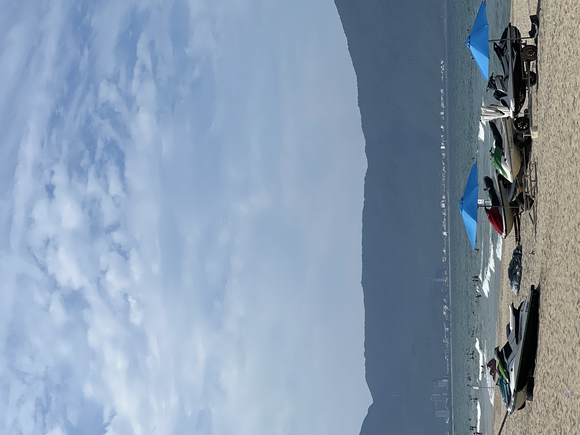 The image captures a scenic view of My Khe Beach in Da Nang, Vietnam, on a bright, overcast day. The foreground is dominated by a sandy beach with several jet skis lined up, some on trailers. Two blue umbrellas provide shade over some of the jet skis. In the middle ground, the ocean is active with small waves and numerous people swimming and enjoying water activities. Beyond the ocean, a distant cityscape with buildings and infrastructure is visible, nestled against a backdrop of imposing, dark, forested mountains that form the horizon. The sky above is a soft blue, filled with scattered white clouds. The overall impression is one of a lively beach resort area on a somewhat hazy day.