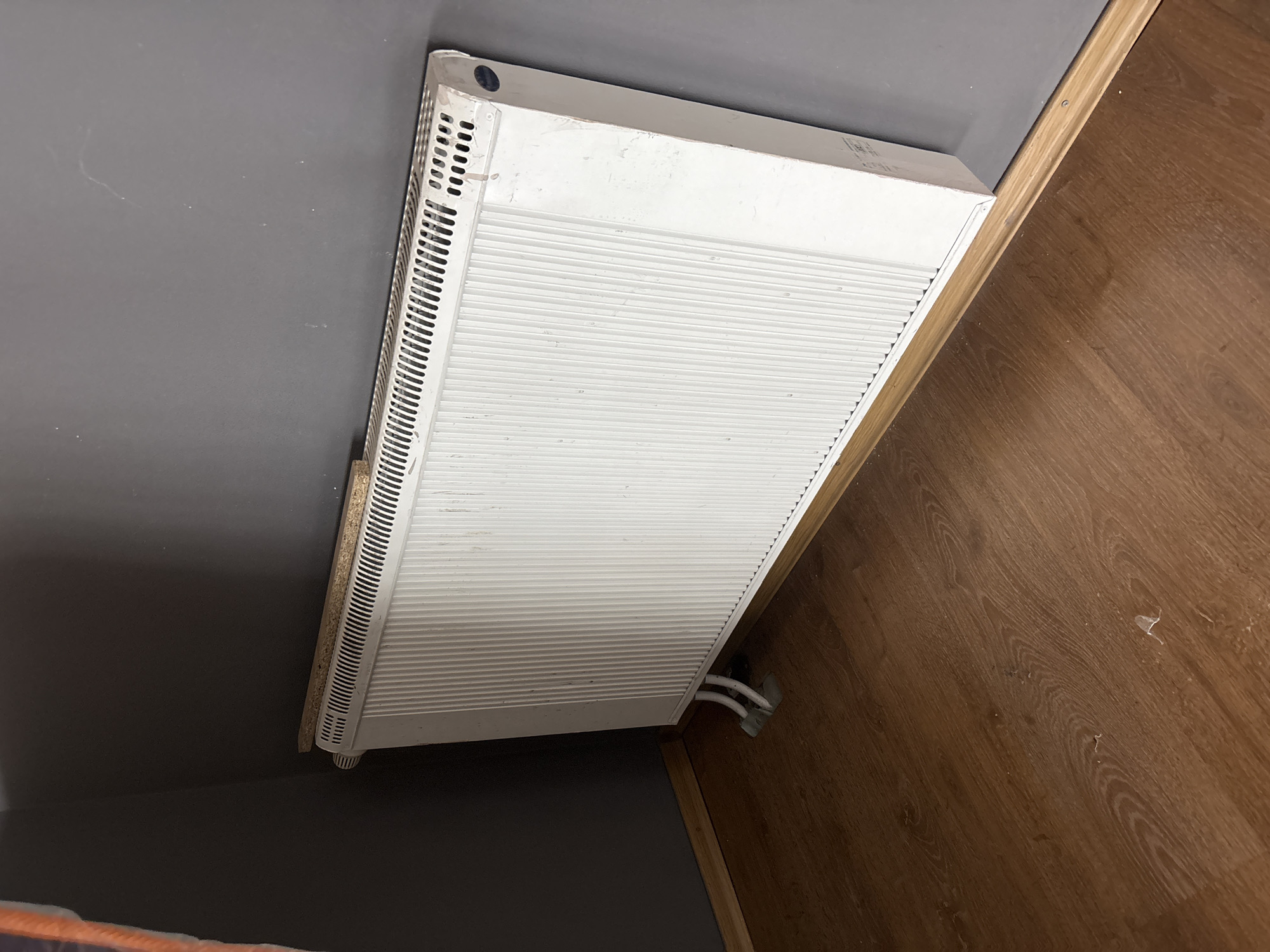 A white radiator stands in the corner of a room, against a grey wall. A wooden plank rests on top of the radiator. The floor is made of brown wood laminate. Two white pipes connect to the radiator at the bottom.