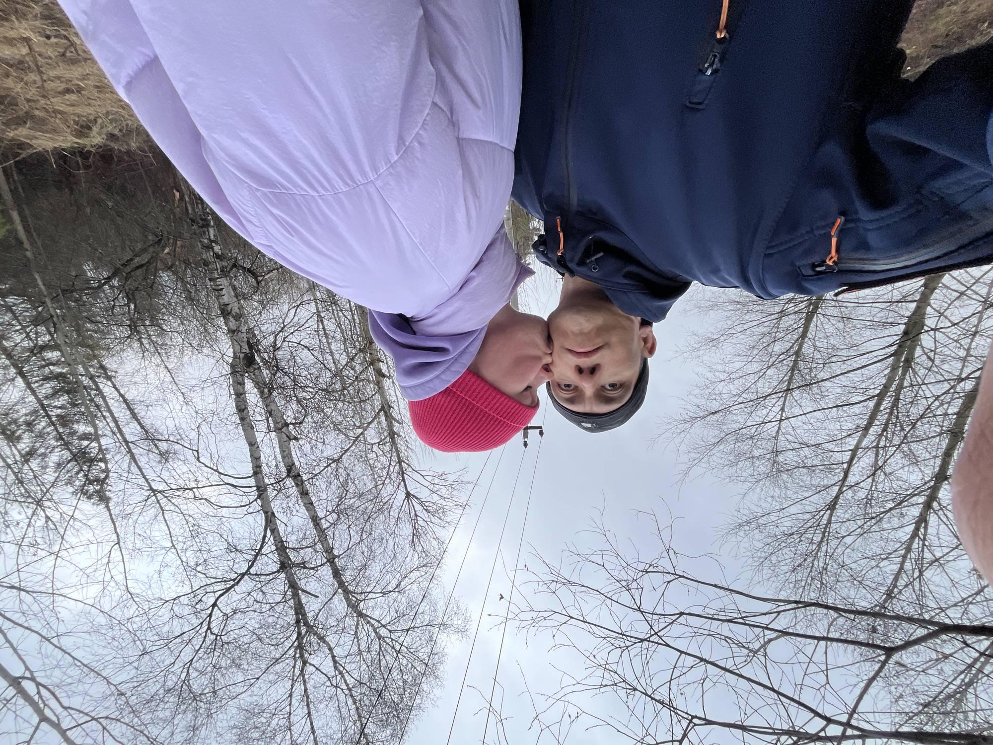 Two individuals are featured prominently at the top of the frame, appearing upside down due to the extreme low-angle perspective and image rotation. One person, on the left, wears a light lavender puffer jacket and a bright pink knitted beanie. They are leaning in, seemingly kissing or nuzzling the cheek of the other person. The second individual, on the right, wears a dark blue jacket with orange zipper accents and a dark grey beanie, looking directly at the camera with a neutral expression.
The setting is an outdoor, possibly wooded or forested area. The background consists primarily of a dense network of bare, leafless tree branches against an overcast, grey sky. Some light-barked trees, possibly birches, are visible. In the middle distance, utility poles and power lines cut across the frame diagonally. The ground, visible towards the "top" of the image (which would be the bottom if the image were righted), shows dry, brownish grass or foliage, suggesting a colder season like late autumn, winter, or early spring. The heavy clothing worn by the individuals supports the idea of cold weather. The image captures a moment of affection between the two individuals, taken from a very unique, upward-looking perspective. There is no visible text.