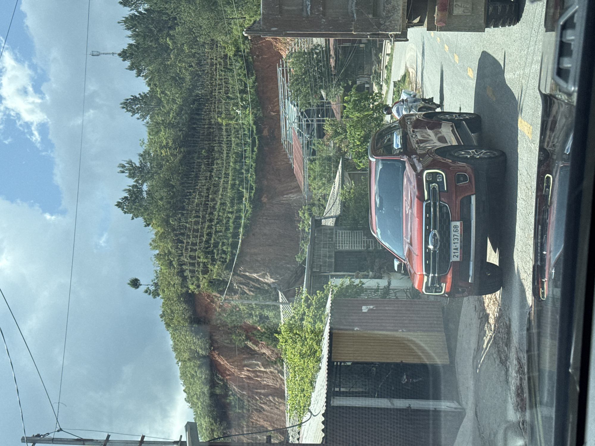 Captured through glass, the image depicts a street scene in Ngã Ba Kim, Vietnam. A red Ford pickup truck, bearing license plate 21A-137.68, is parked on the right side of the road. Adjacent to the truck is a motorcycle with two people on it. A large truck is positioned ahead of the Ford. In the background, a hillside densely covered with vegetation is evident, supported by a retaining wall. A utility pole is visible in the distance against a cloudy sky. Buildings and scaffolding are also present. The photograph appears to be taken from inside a vehicle.