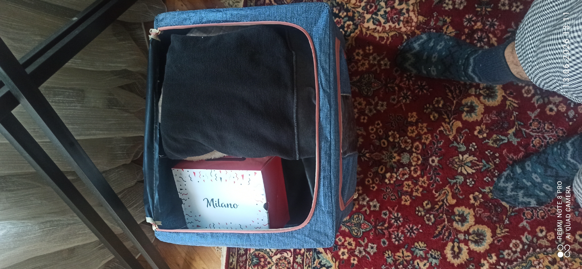 An overhead shot shows a person's feet wearing patterned socks and plaid pants on a red oriental rug. To the right, a foot is visible wearing a dark blue sock with a white zig-zag pattern. Above the rug is a blue fabric storage cube with a pink trim. Inside the cube, a white box with "Milano" written in cursive on it is visible. Next to the box, a folded piece of dark fabric, possibly clothing, is placed inside the cube. The background is a blurred view of sheer curtains.