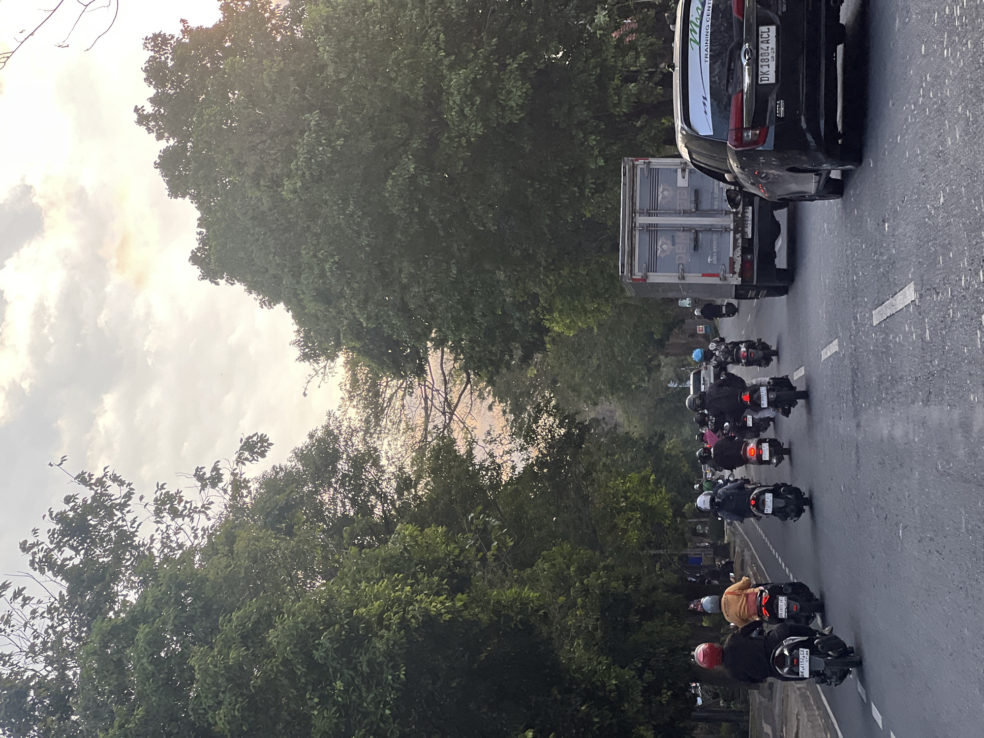 The image depicts a street scene in Denpasar, Indonesia, likely during the late afternoon or early evening. The road is lined with lush, mature trees on both sides, creating a canopy over the asphalt. A group of motorcyclists, predominantly wearing helmets, are riding in the same direction, with some in the foreground and others further down the road. A black SUV is in the right lane, displaying a license plate with "DK 1884 ACL." A delivery truck with "DBM" branding is ahead of the SUV. The sky above is partly cloudy with warm, diffused sunlight suggesting either sunrise or sunset. The atmosphere is one of moderate traffic flow on a tree-lined urban street.
