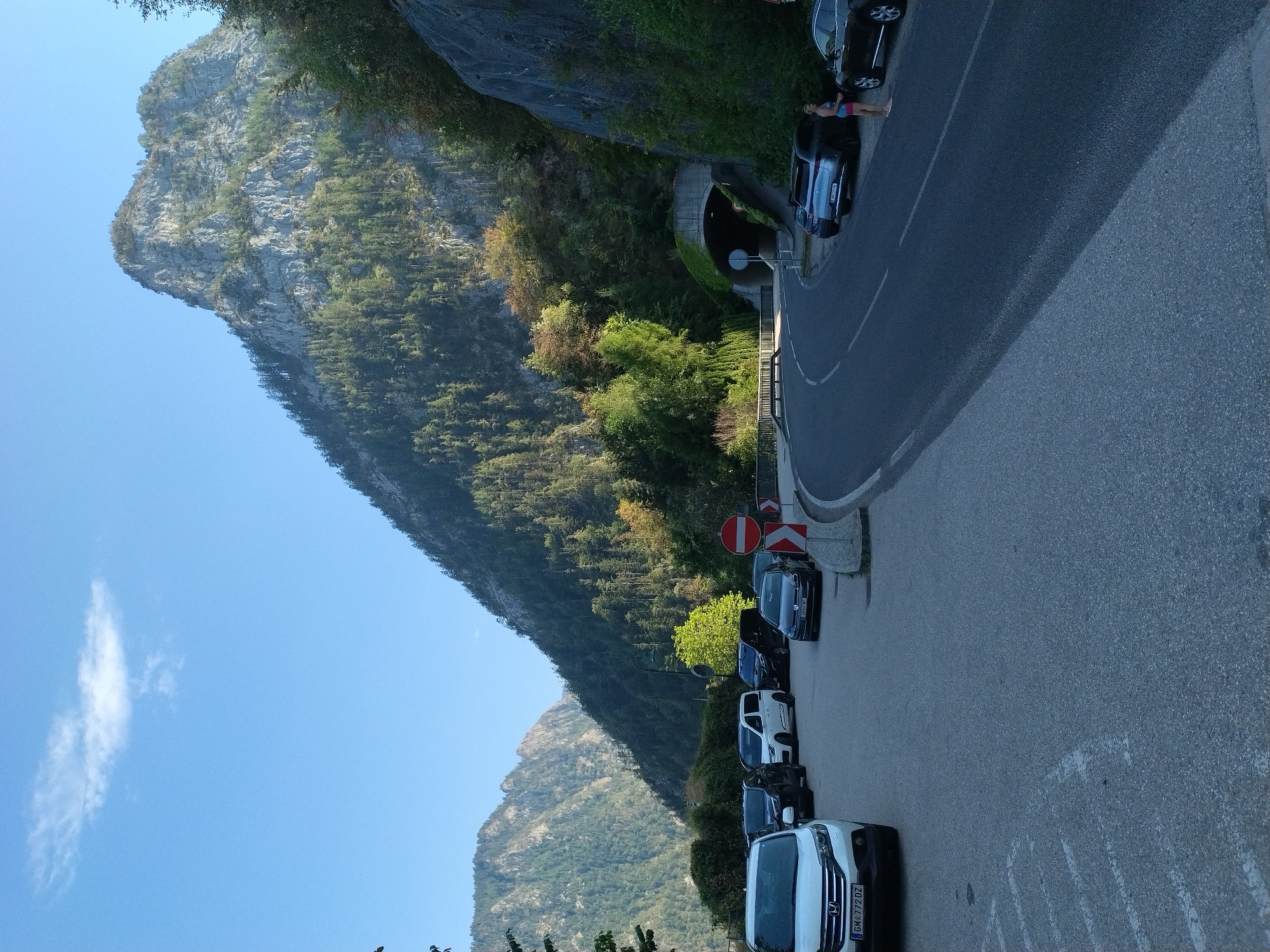 The image depicts a scenic view of a road curving around a mountainside in Traunkirchen, Austria, leading towards a tunnel entrance. The road is lined with parked cars on the left side, including a white SUV with the license plate "GM 772 DZ", several dark-colored sedans, and a white minivan. Further down the road, a woman in athletic wear is standing near a parked dark SUV. The mountainside is densely covered with evergreen trees, and a prominent rocky peak dominates the upper right portion of the frame. A single, thin cloud is visible in the bright blue sky. The environment appears to be a natural, possibly tourist-oriented area, with a tunnel indicating a passage through the mountain.