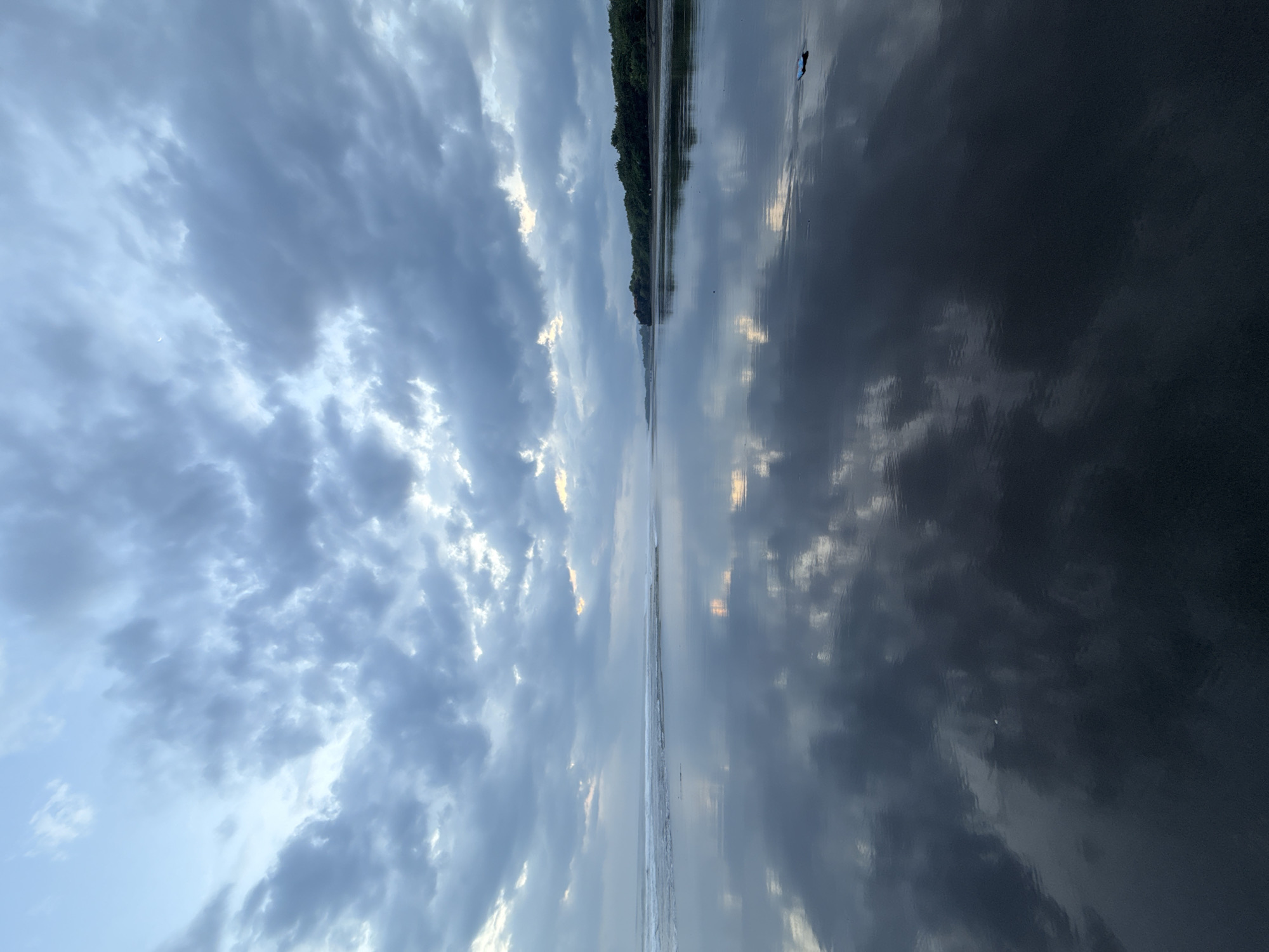The image depicts a body of water, possibly a river or lake, reflecting a sky filled with gray and white clouds. A thin strip of land with trees is visible on the horizon, dividing the sky from the water. The scene is bisected vertically, showing the reflection of the sky in the water. The location is stated as Unknown, Unknown.